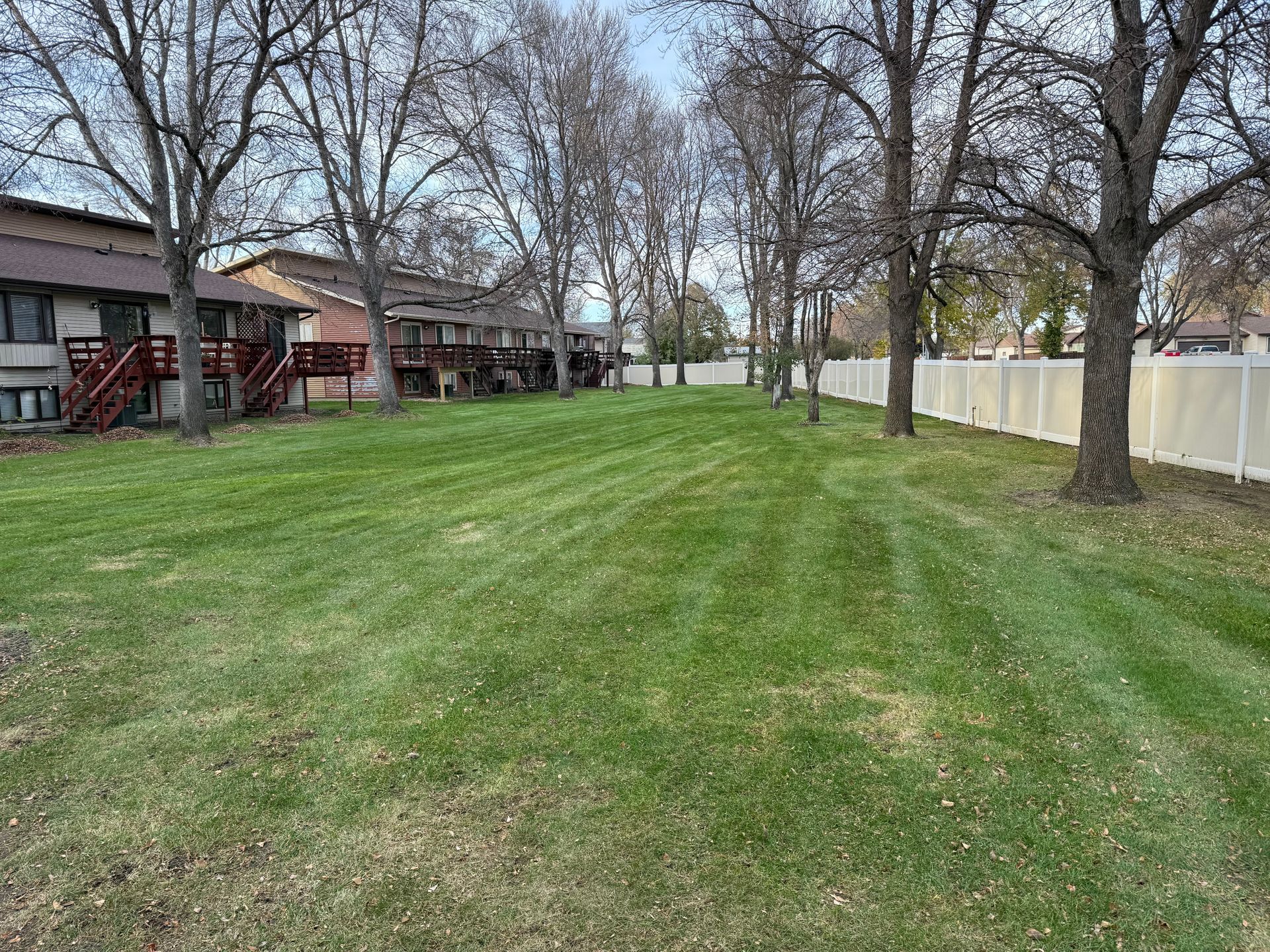 Green lawn with apartment buildings on the left and a white fence on the right. Trees line the right side.