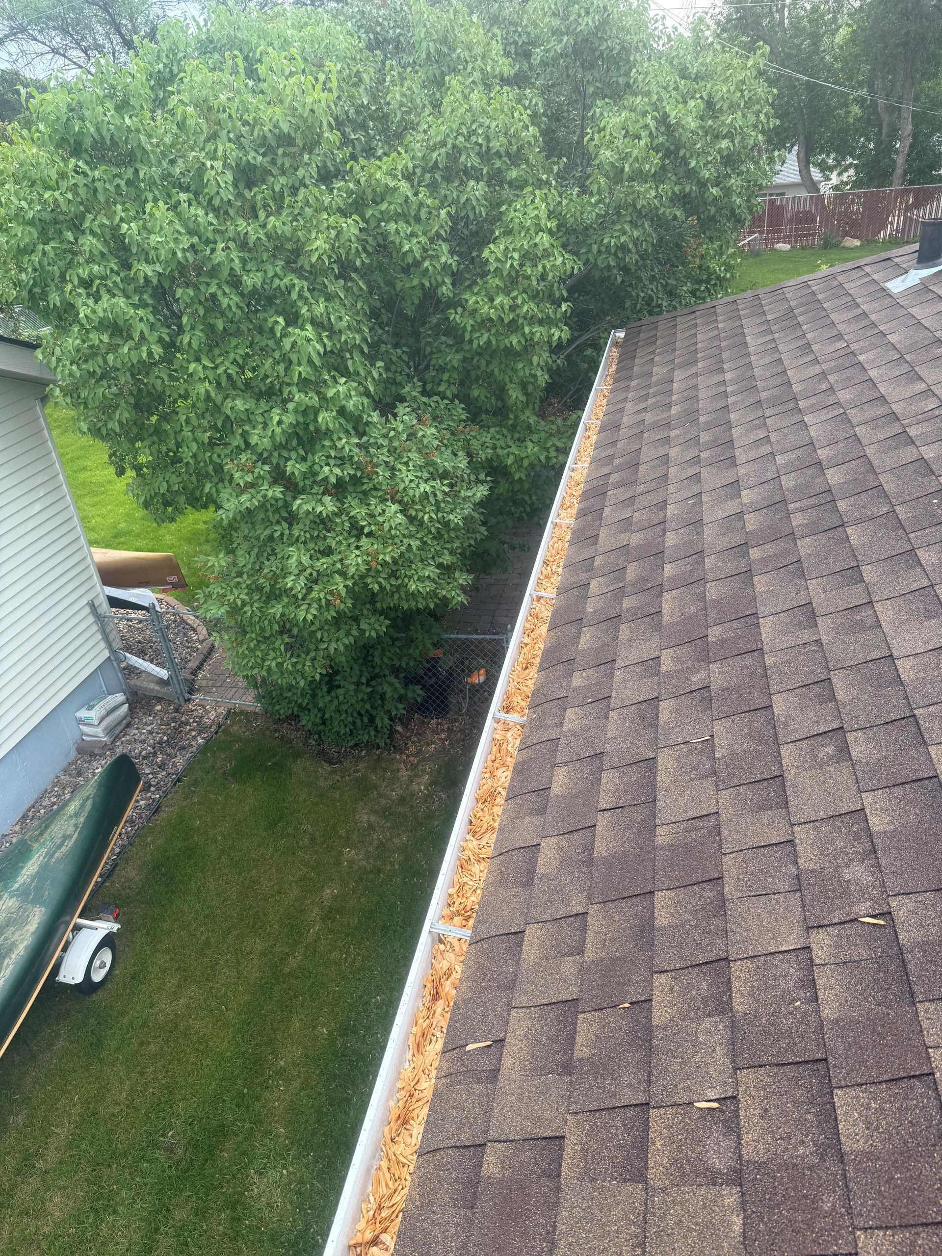 Roof with brown shingles, gutter with debris, and green trees.