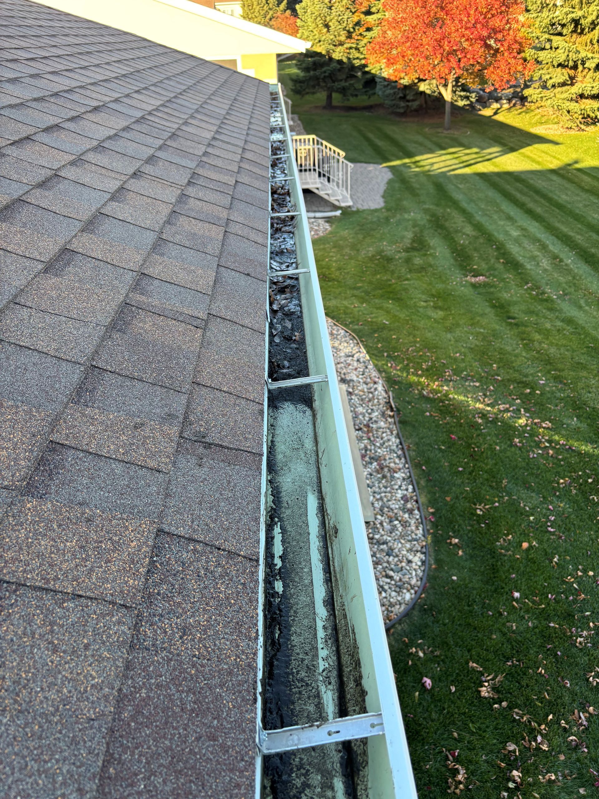 Gutter filled with debris next to a shingled roof, with green grass and fall foliage in the background.