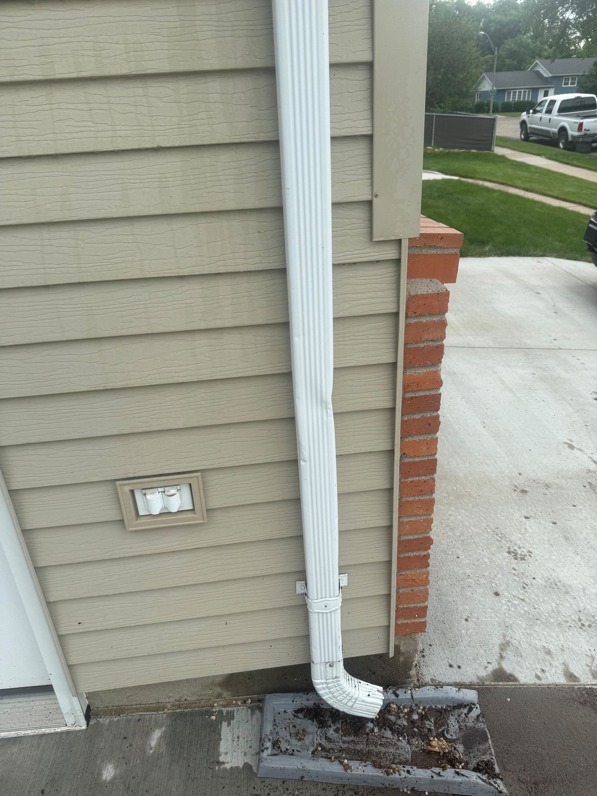 White guttering against beige siding and brick column, leads to ground level downspout.