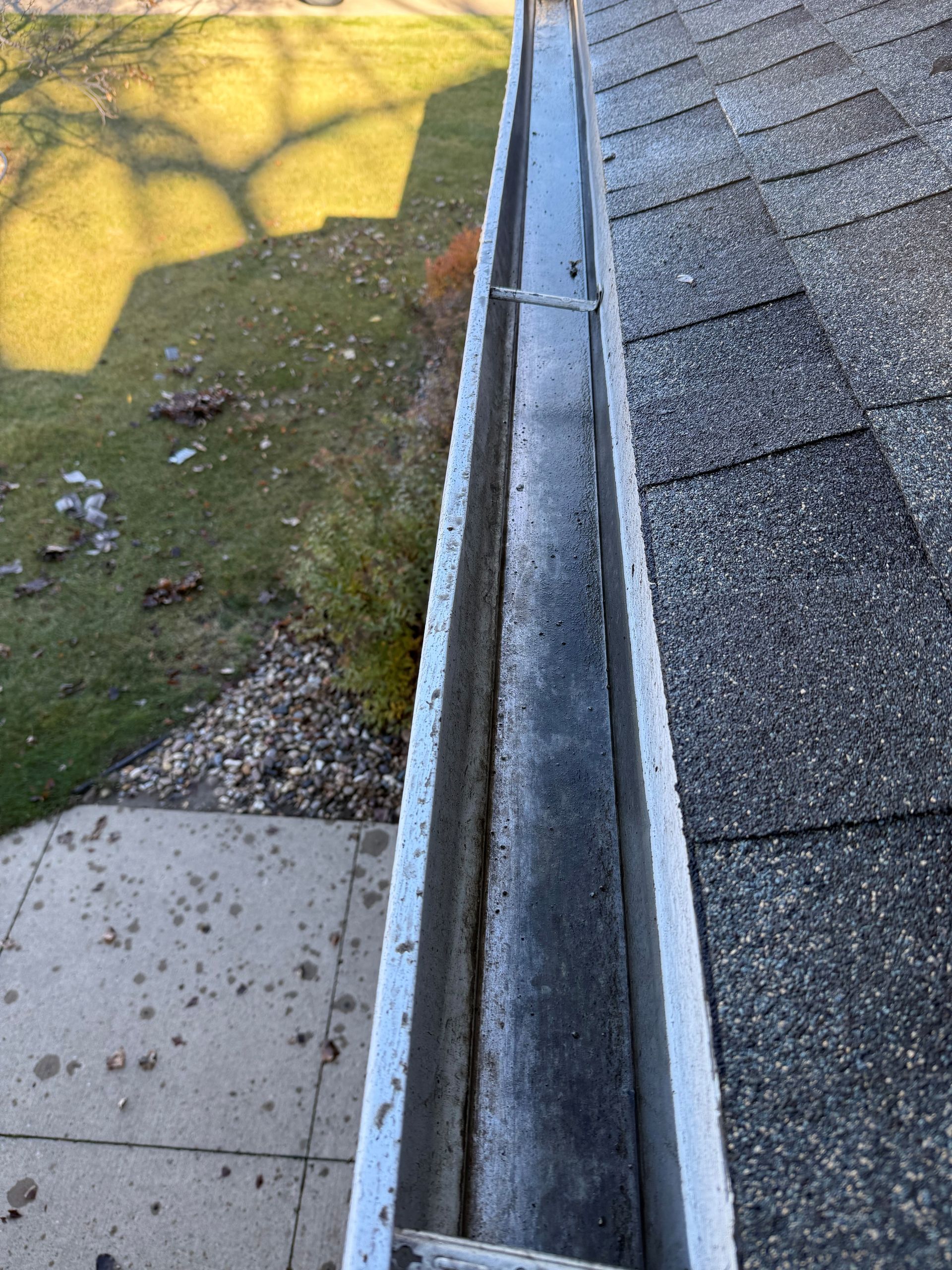 Gutter on a roof with visible debris, viewed from above, with grass and a concrete path in the background.