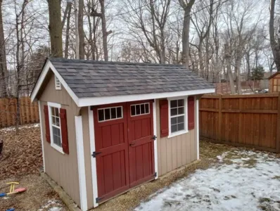 A small shed with red doors and shutters in a snowy backyard.