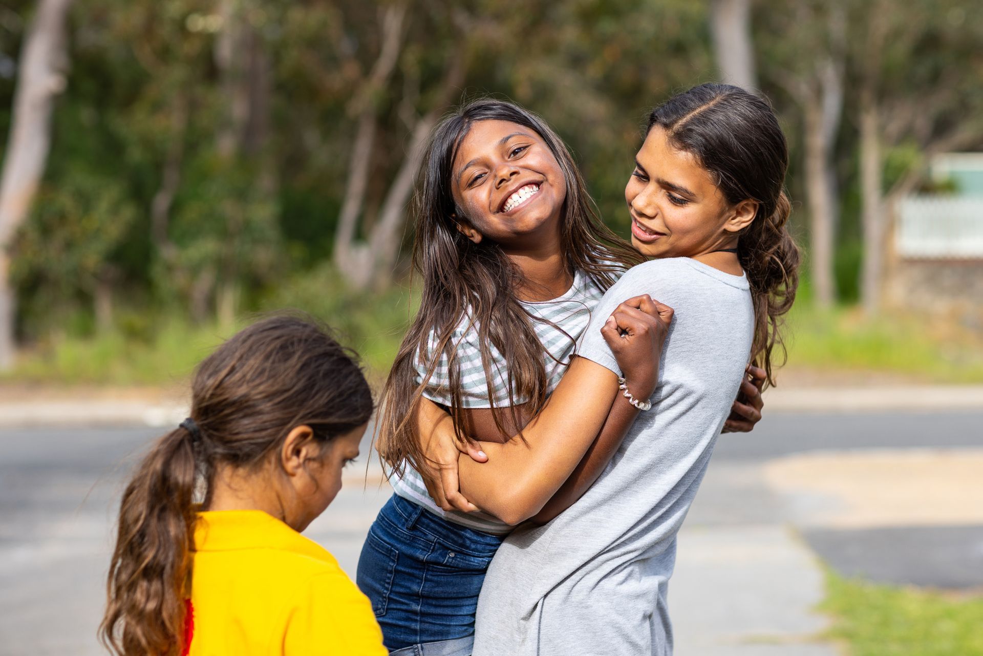Two smiling children hugging outdoors, with another child in a yellow shirt nearby.