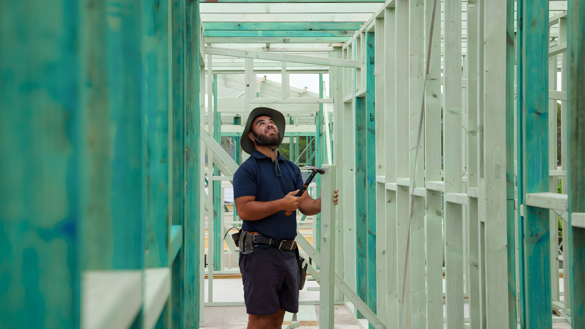 Worker in blue shirt and safety harness stands in a teal-framed construction corridor, looking up.