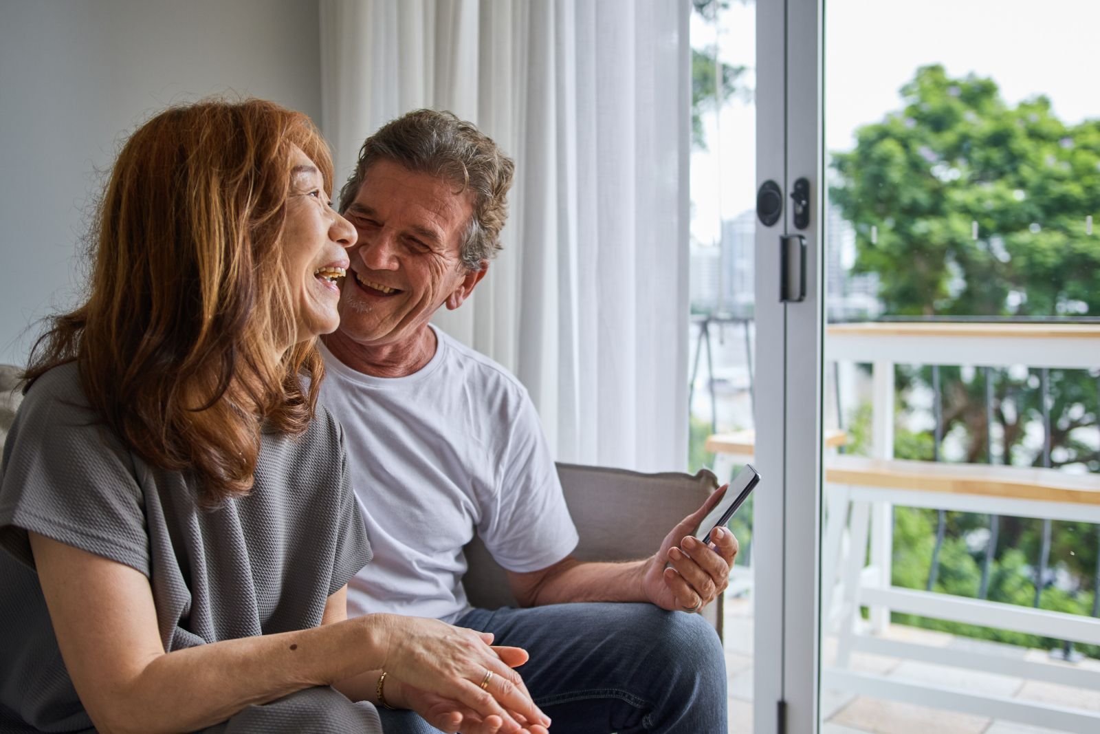 Two people sit by a bright window, smiling and looking at a smartphone together.
