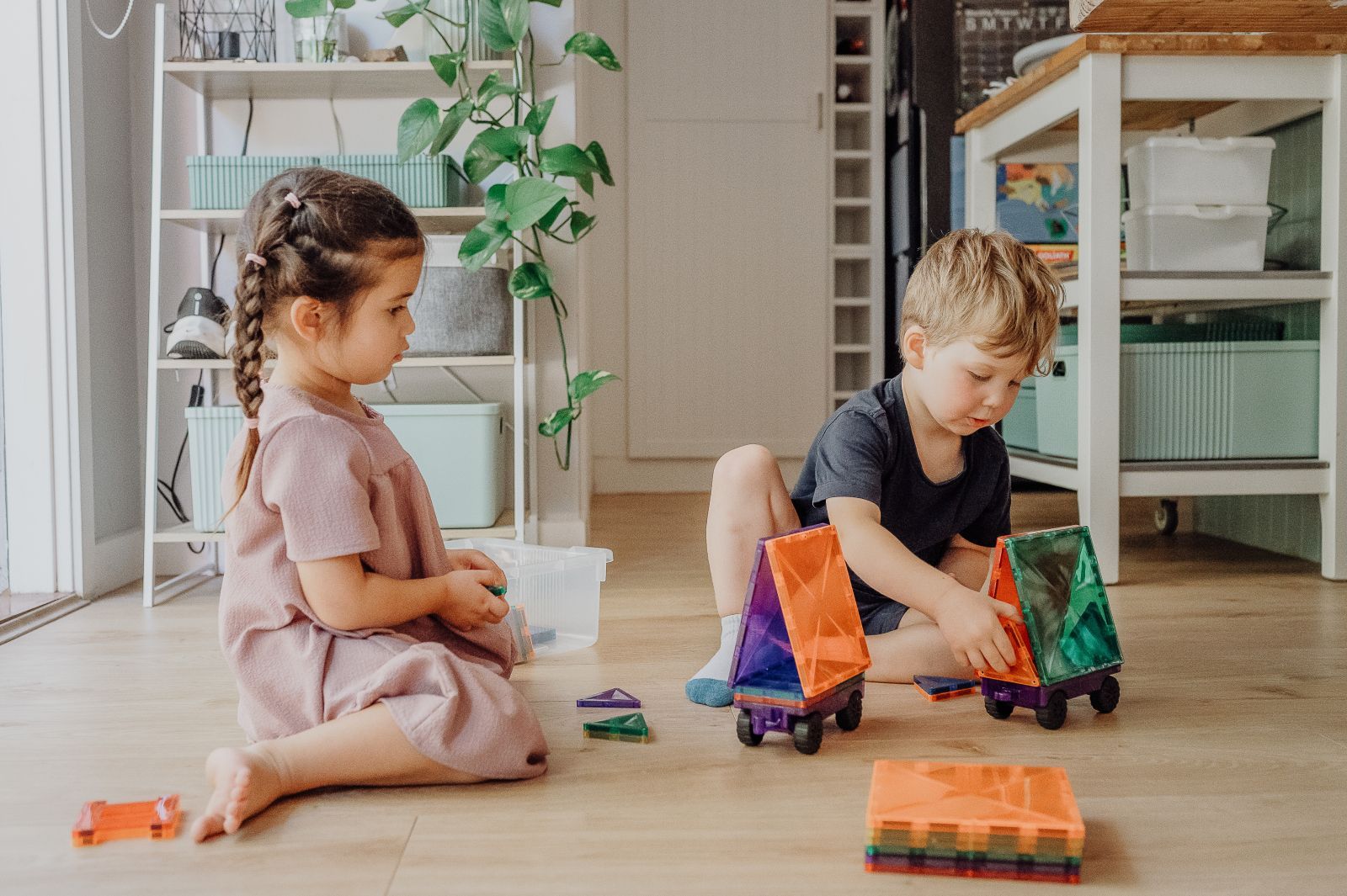 Two children playing with toy cars on a wooden floor in a bright room