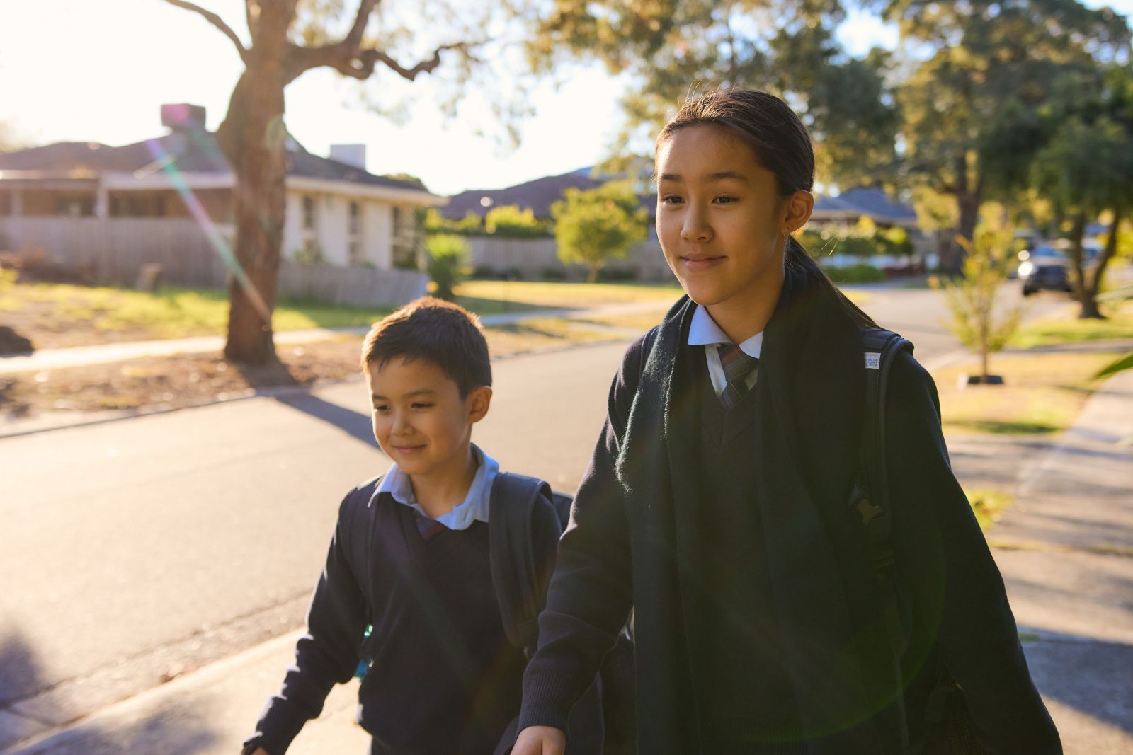 Two children walking on a sunny suburban street, one smiling, backpacks on, with houses and trees in the background