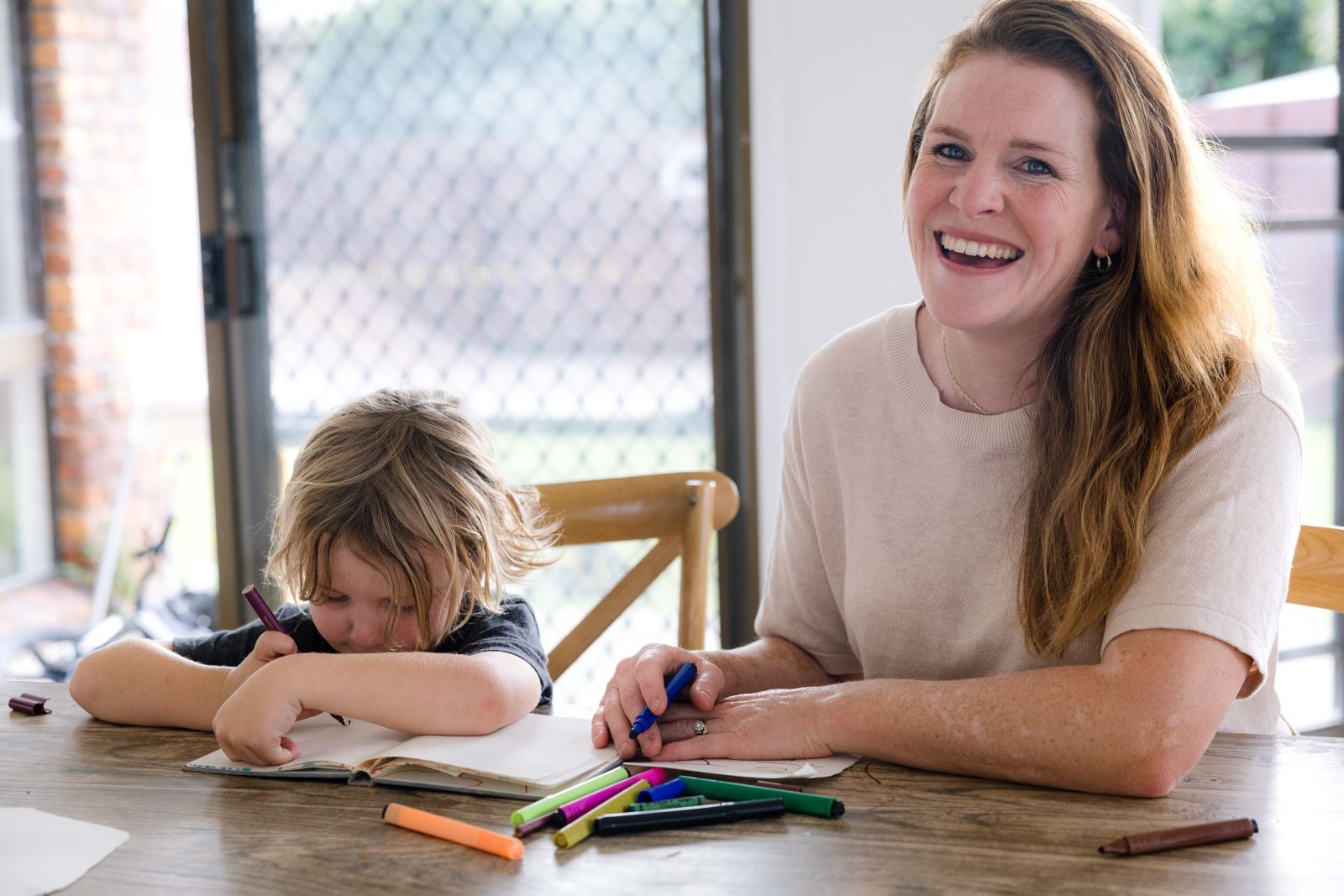 Woman smiling beside child drawing at a table with colored pencils in a bright room