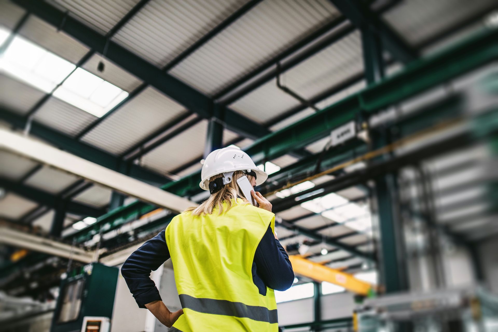 Woman in safety vest and hard hat talks on the phone in a factory.