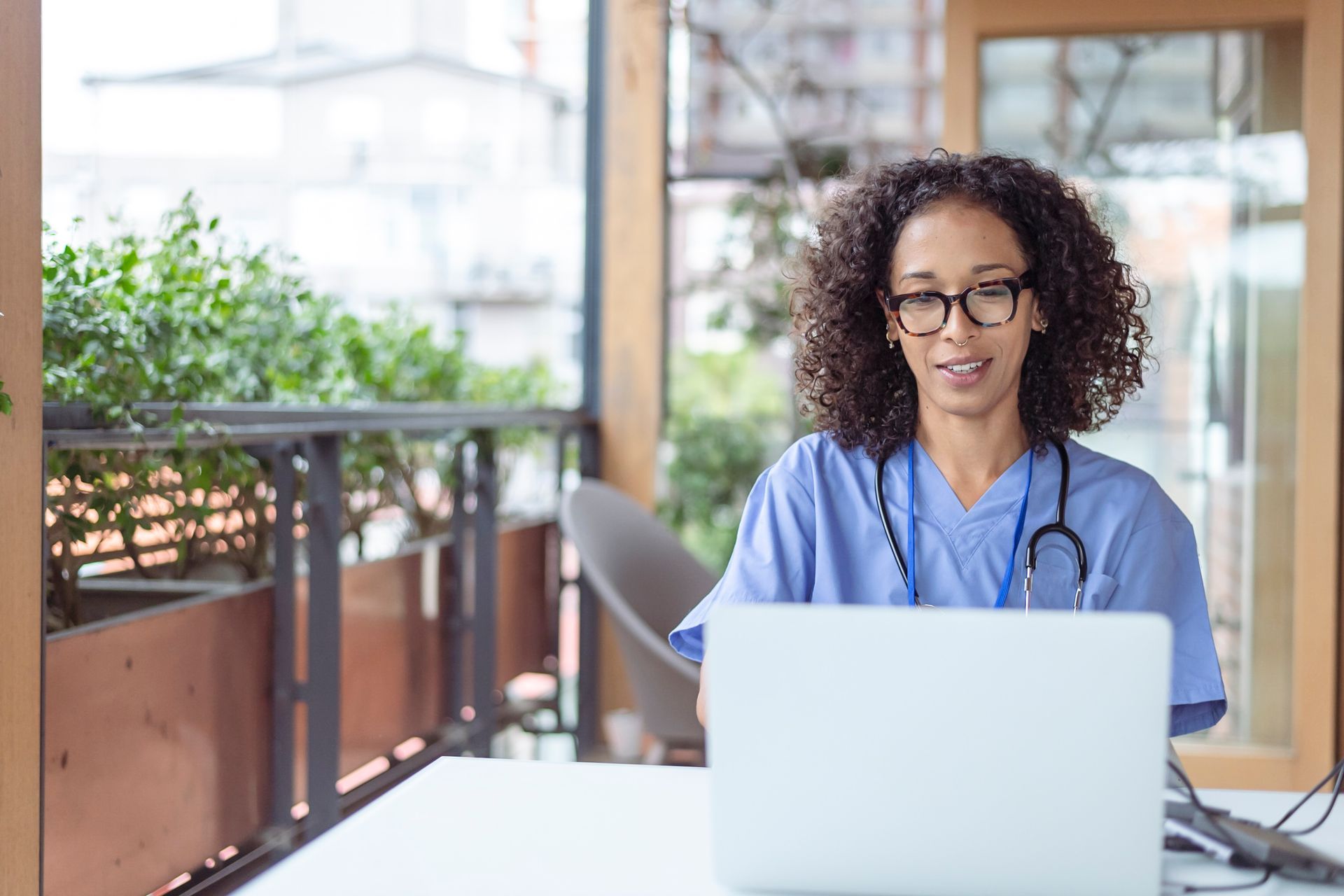 Healthcare worker in blue scrubs smiling at a laptop on a bright outdoor patio
