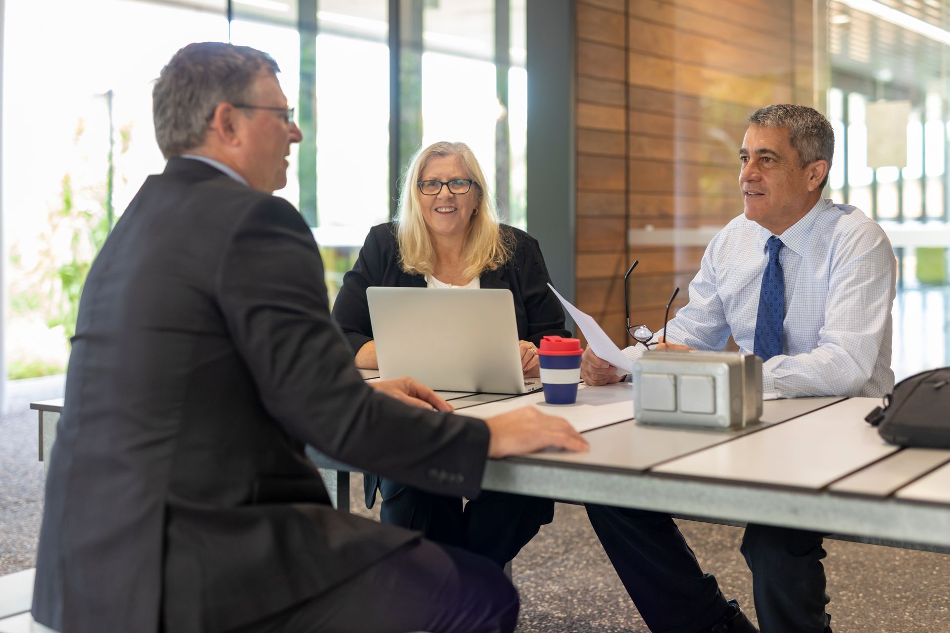 Three people in business attire meeting at an outdoor table with a laptop and papers