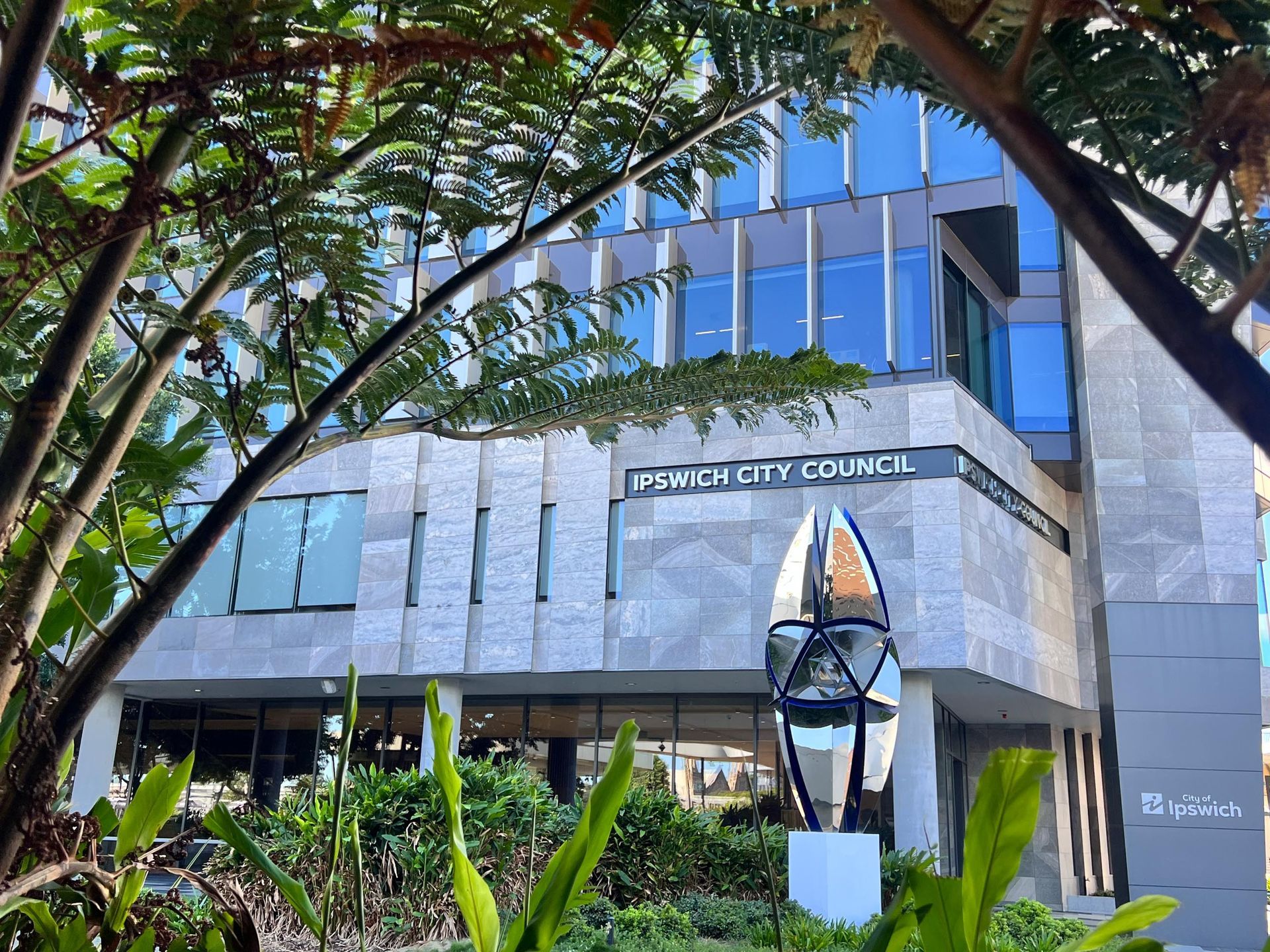 Modern glass office building with a sign, framed by trees and greenery