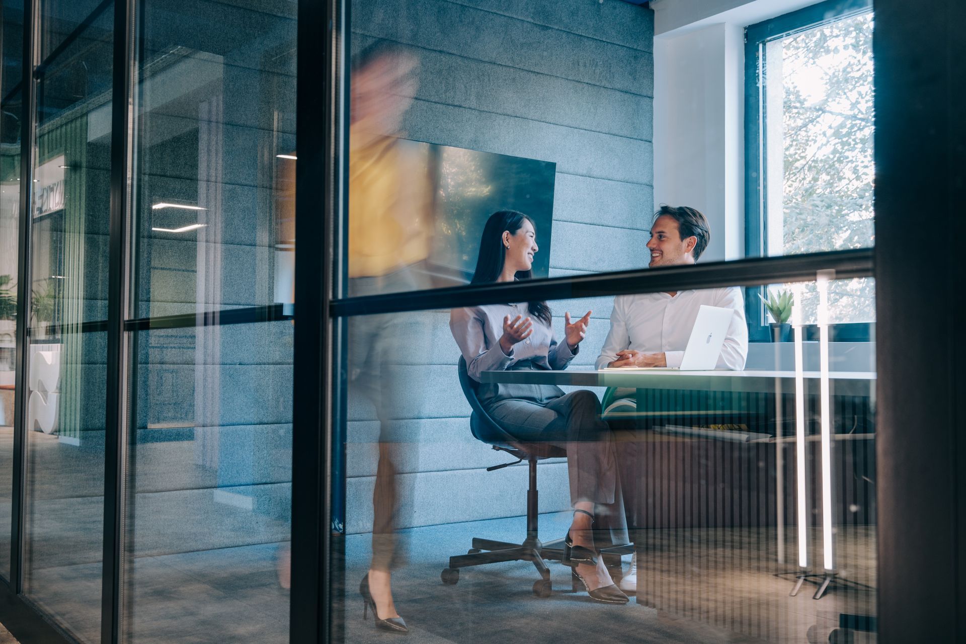 Glass-walled office meeting with two people talking at a table, seen through a hallway window.