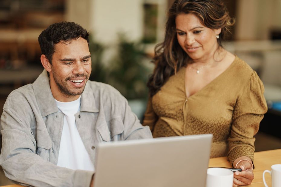 Two coworkers smiling over a laptop at a table, with coffee cups nearby