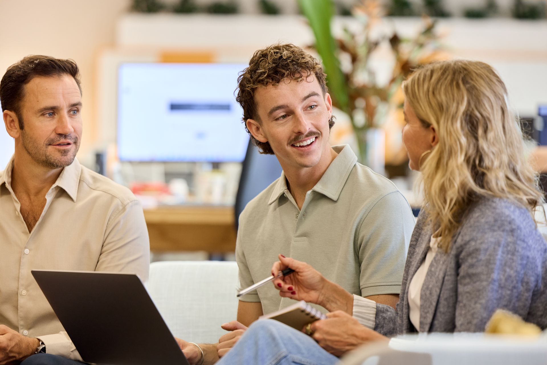 Three coworkers discussing a laptop in a bright office, smiling and gesturing during a meeting