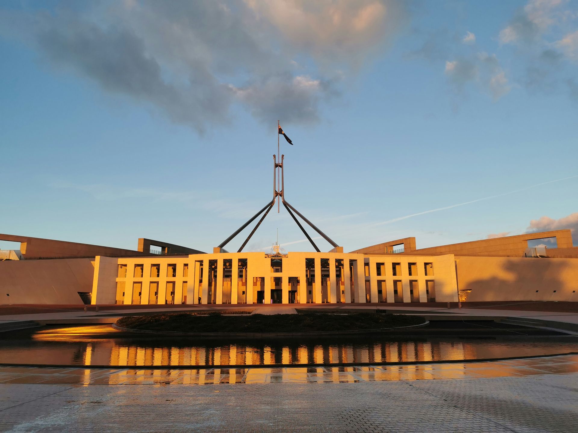 Parliament building at sunset, with a flagpole and reflections in the foreground water.