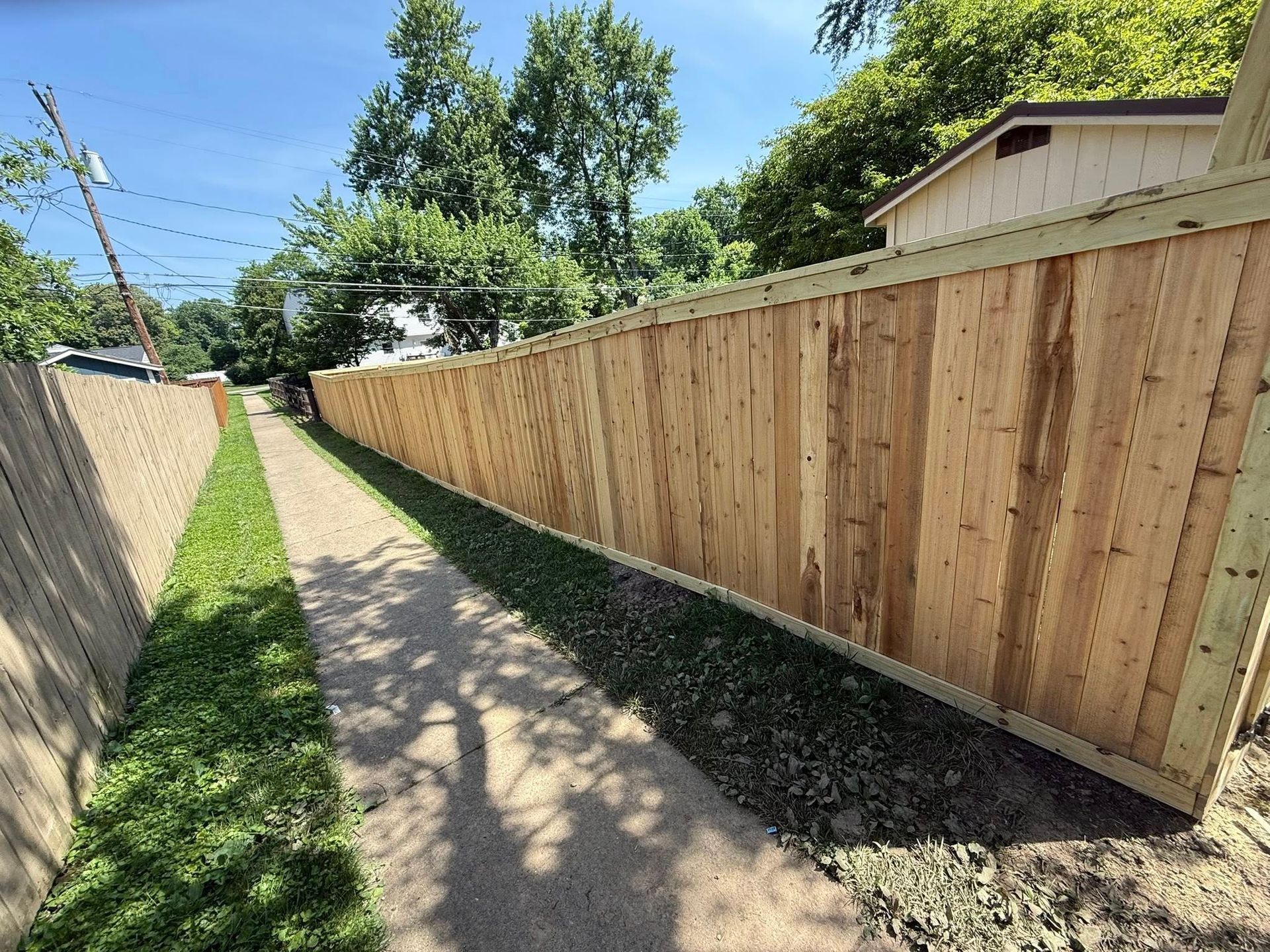 A narrow gravel path flanked by tall wooden fences and grass under a bright blue sky.
