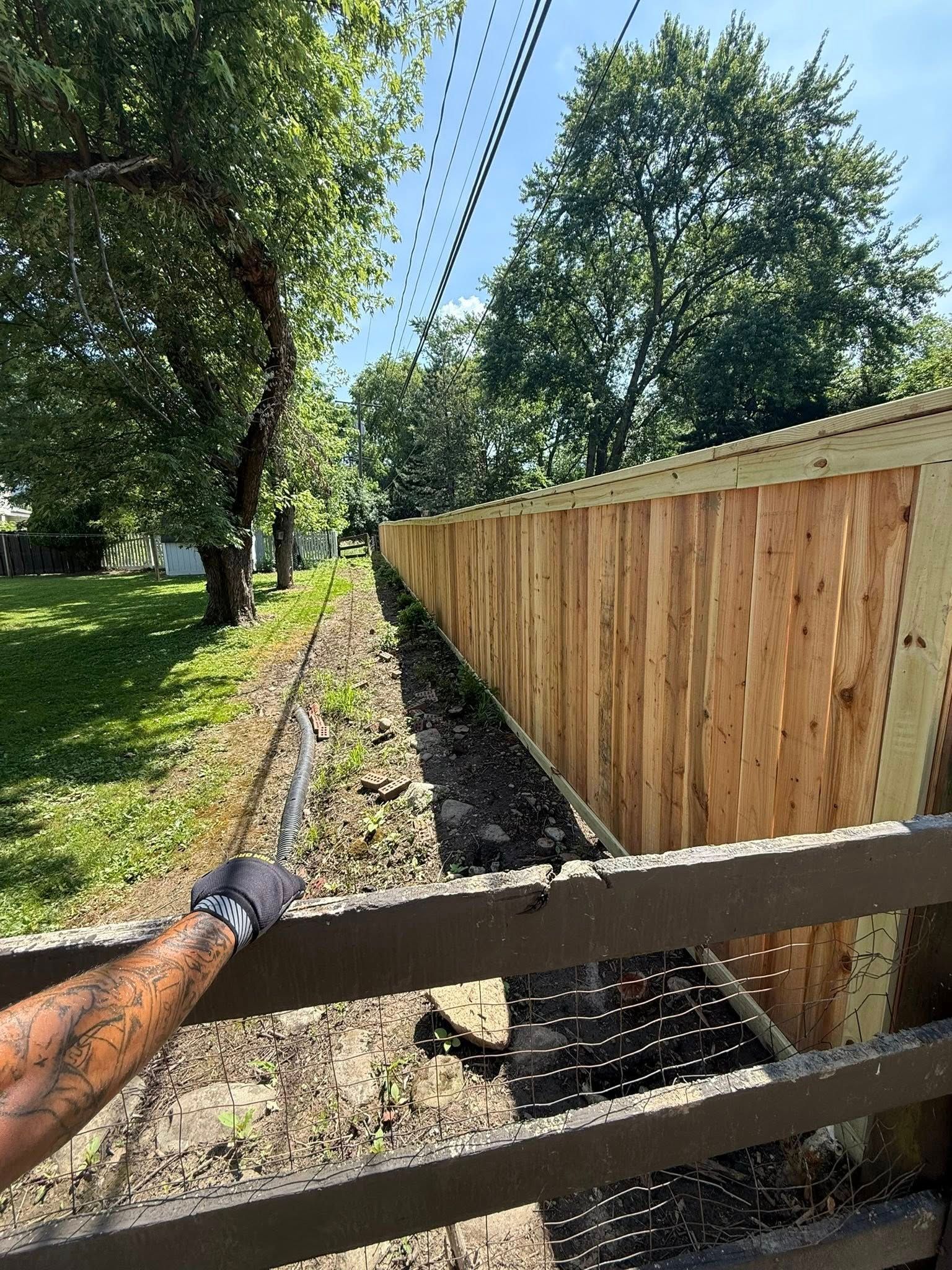 A person with arm tattoos holds a tool, looking towards a newly built wooden fence next to a grassy area.
