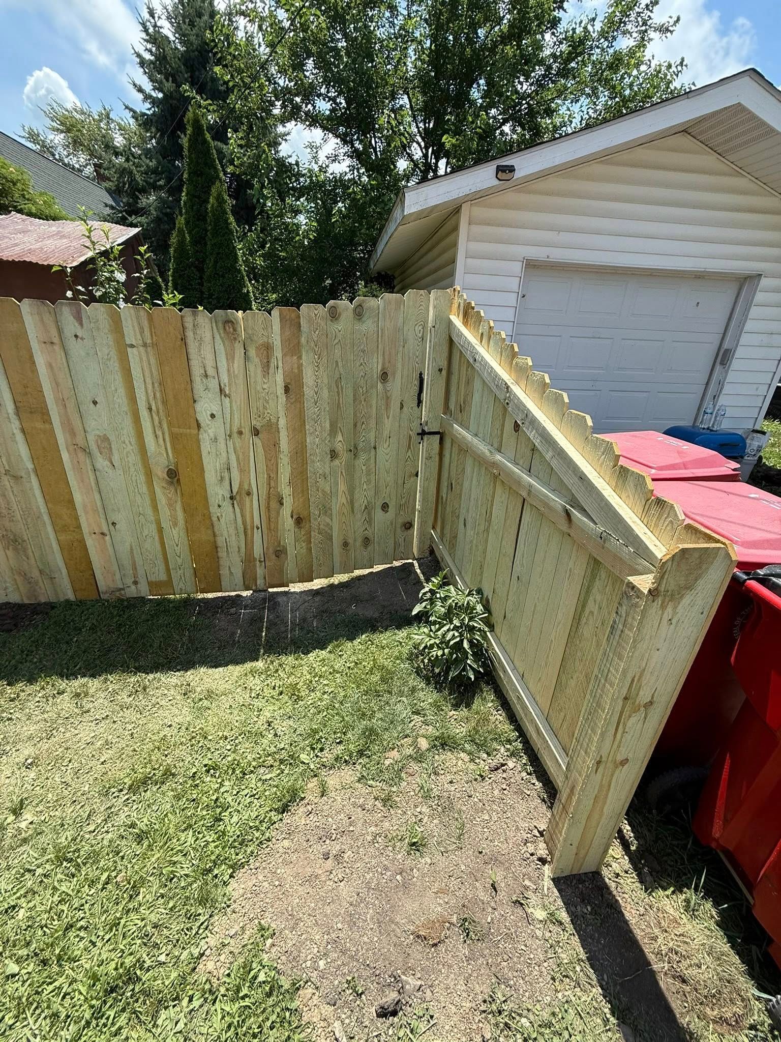 Wooden fence in backyard, adjacent to garage. Green grass, red trash bin.