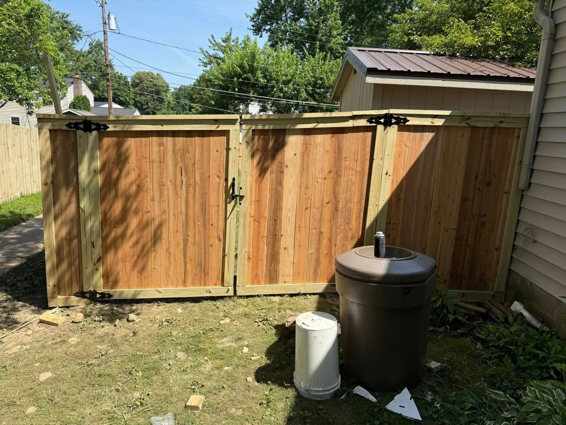 Wooden double gate in a yard; brown trash cans and a white pipe stand in front.