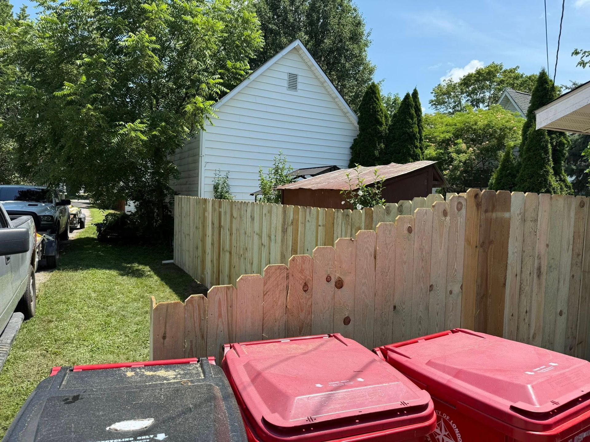 A wooden fence lines a yard with trash cans in front. A white building is in the background.
