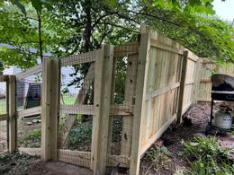 Wooden fence with a gate in a backyard, overgrown with greenery; a grill is visible.