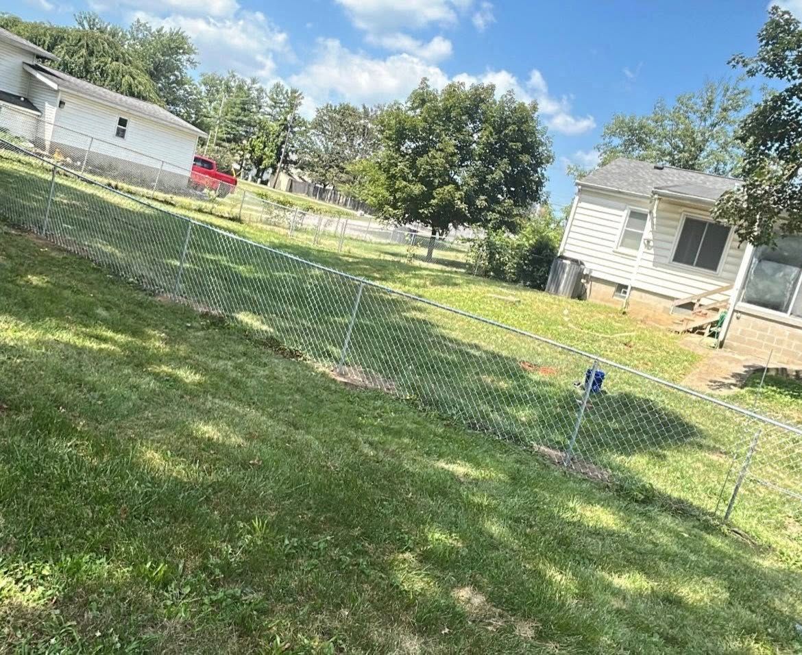 A sloped, grassy yard with a chain-link fence. White houses are on either side, under a blue sky.