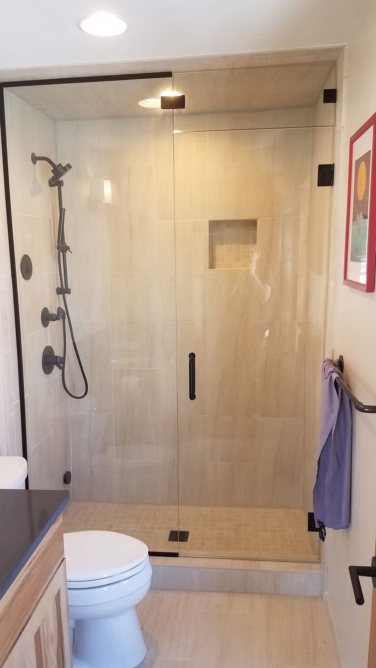 Bathroom with glass-enclosed shower, featuring black fixtures, light-colored tile, and a toilet. A towel hangs on a rack.
