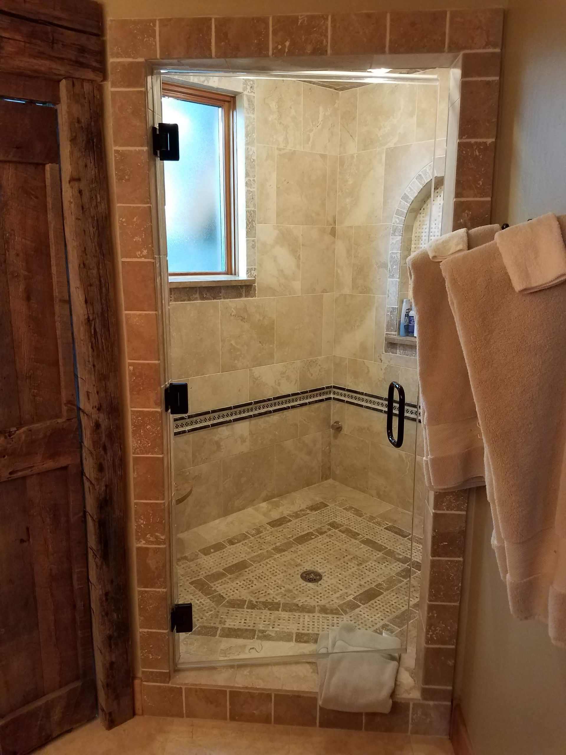 A walk-in shower with a glass door and beige tile. The shower has a small window and is framed by dark brown tile.
