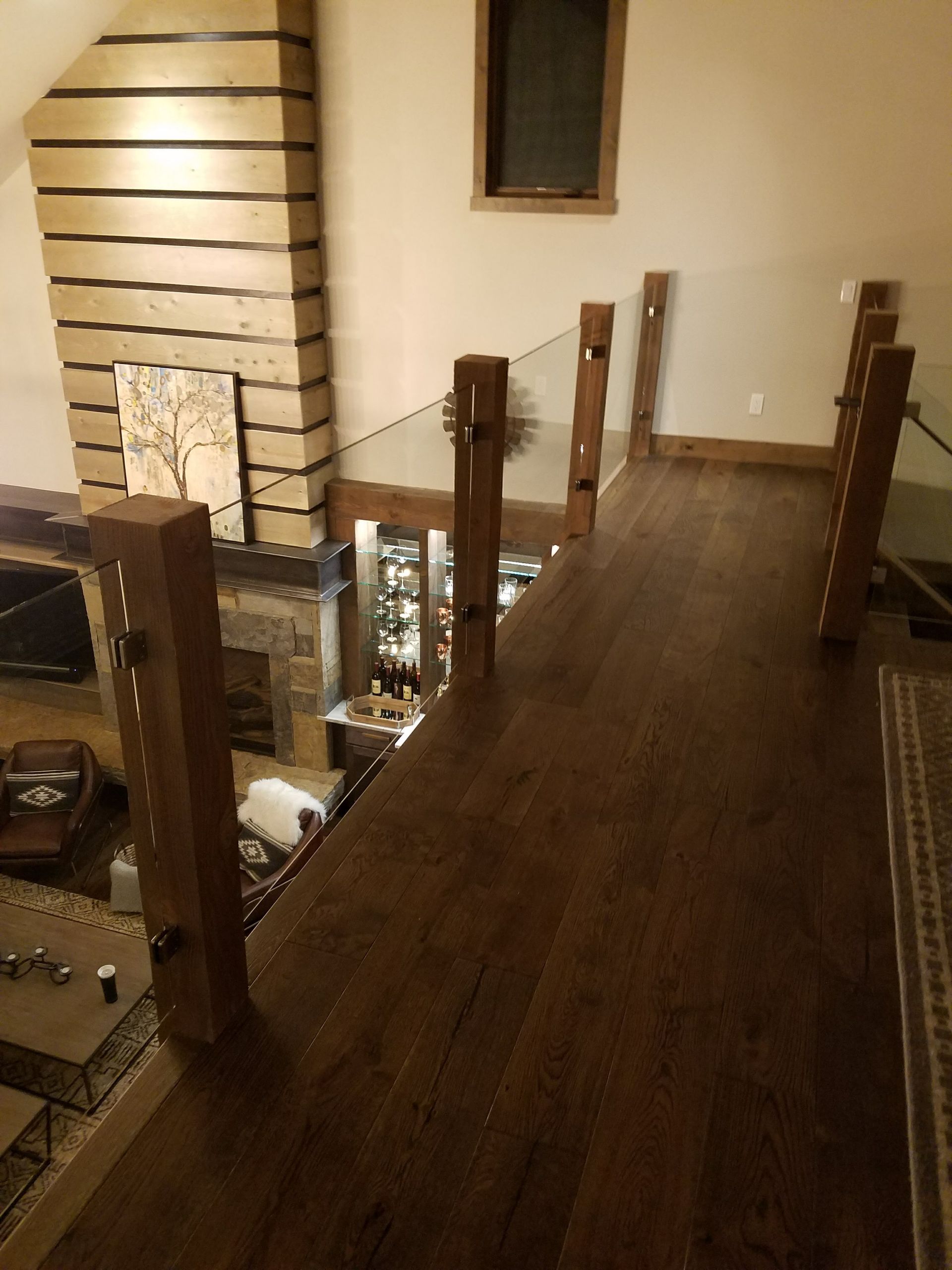 Wooden balcony with glass railing overlooking a living room with fireplace and bar.