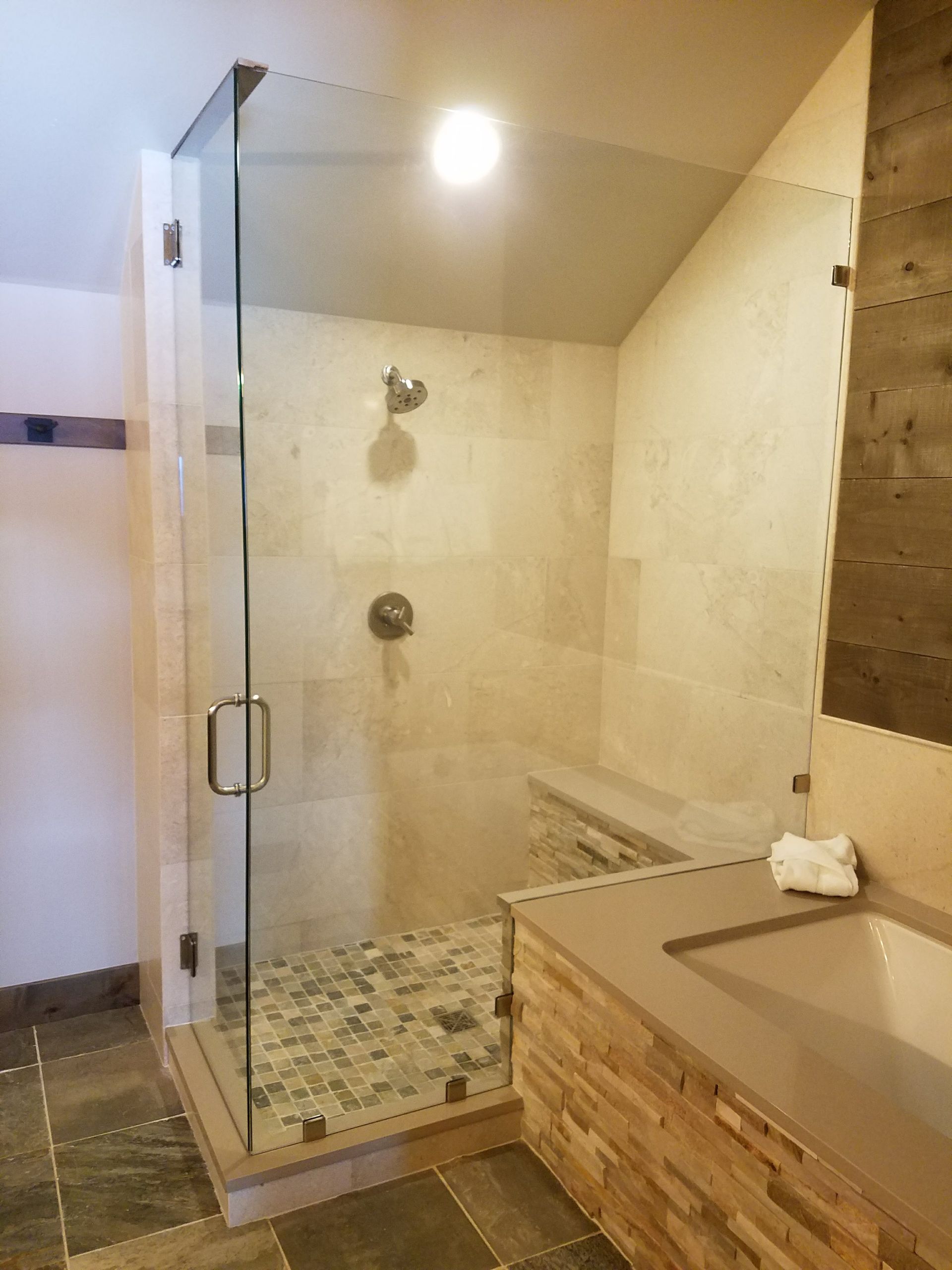 Glass shower enclosure with a stone bench, shower head, and vanity with a stone facade. The walls are tiled.
