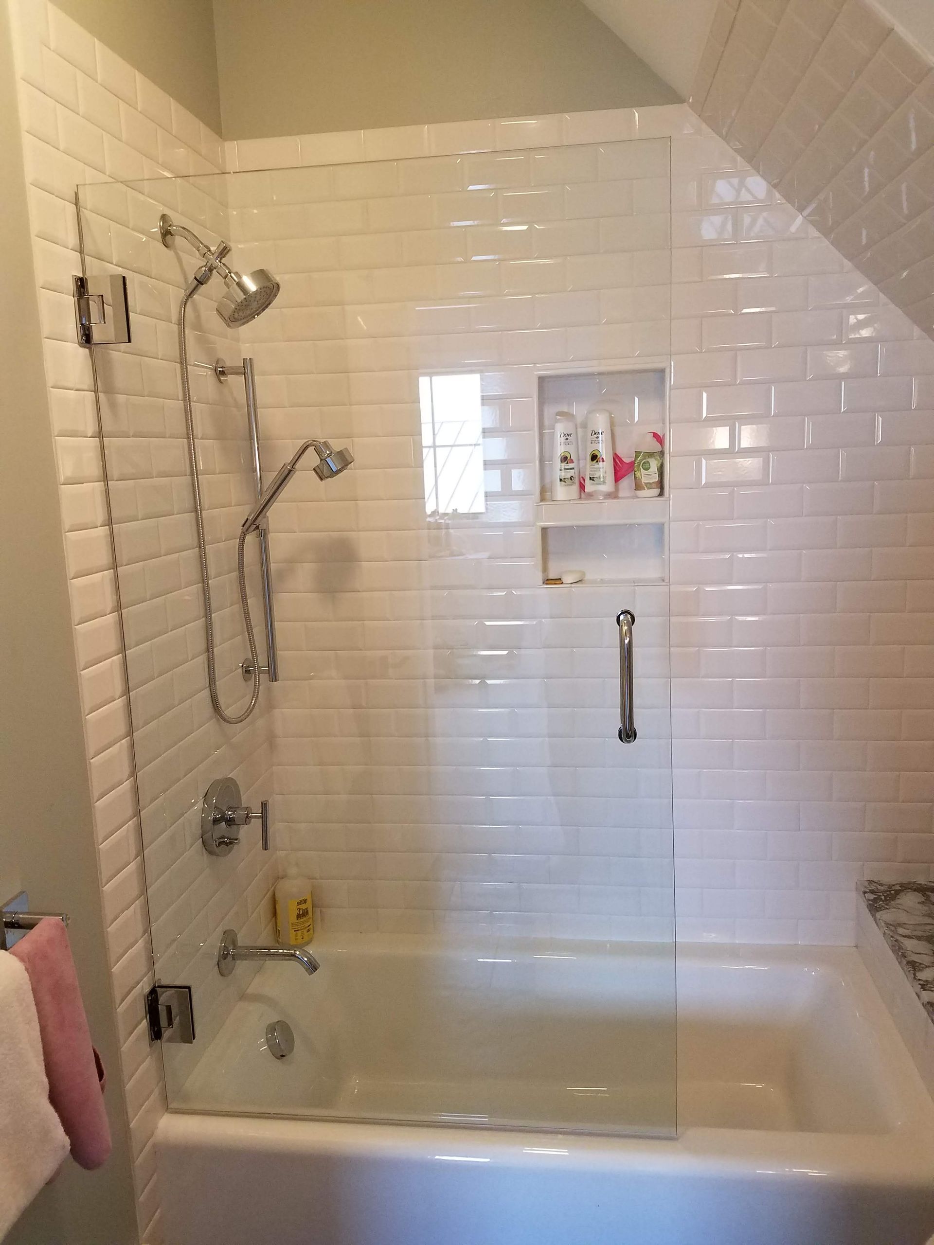 White tiled shower with glass door, built over a bathtub. A shelf with shampoo bottles is built into the wall.