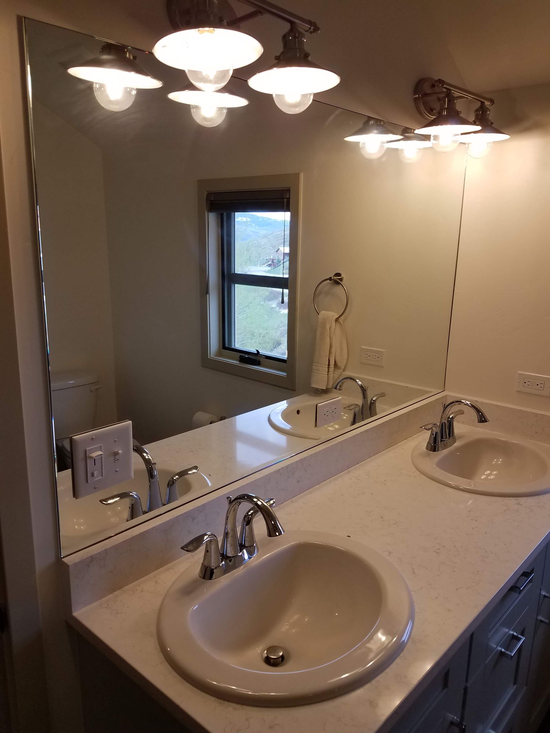 Bathroom with a long mirror, two sinks, and overhead lighting. A window reflects an outdoor scene.