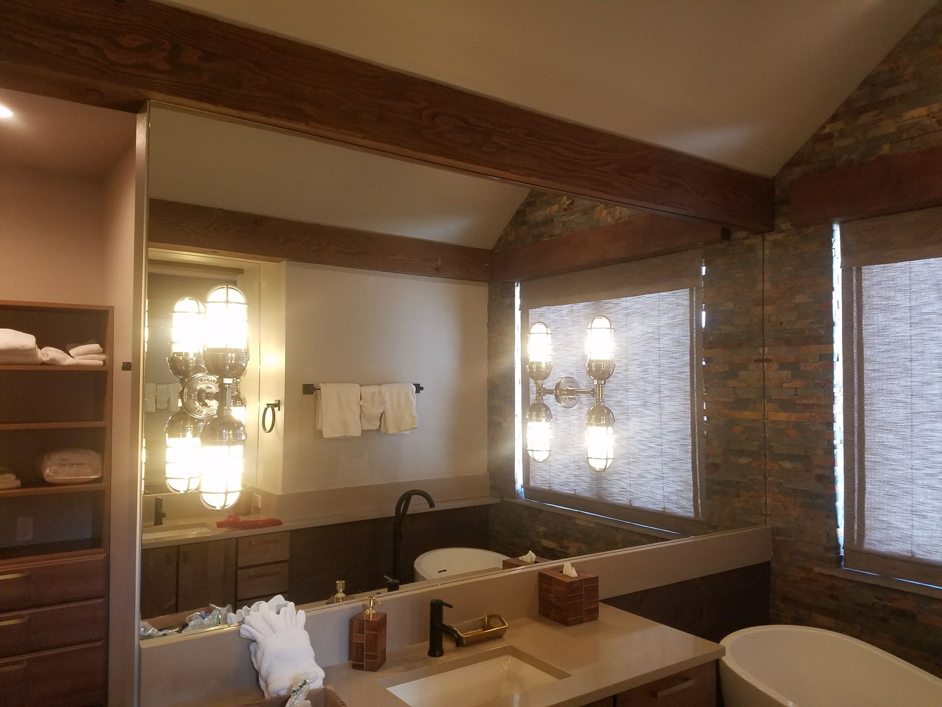 Bathroom with rustic wood beams, stone accent wall, large mirror, vanity, and a freestanding tub.  