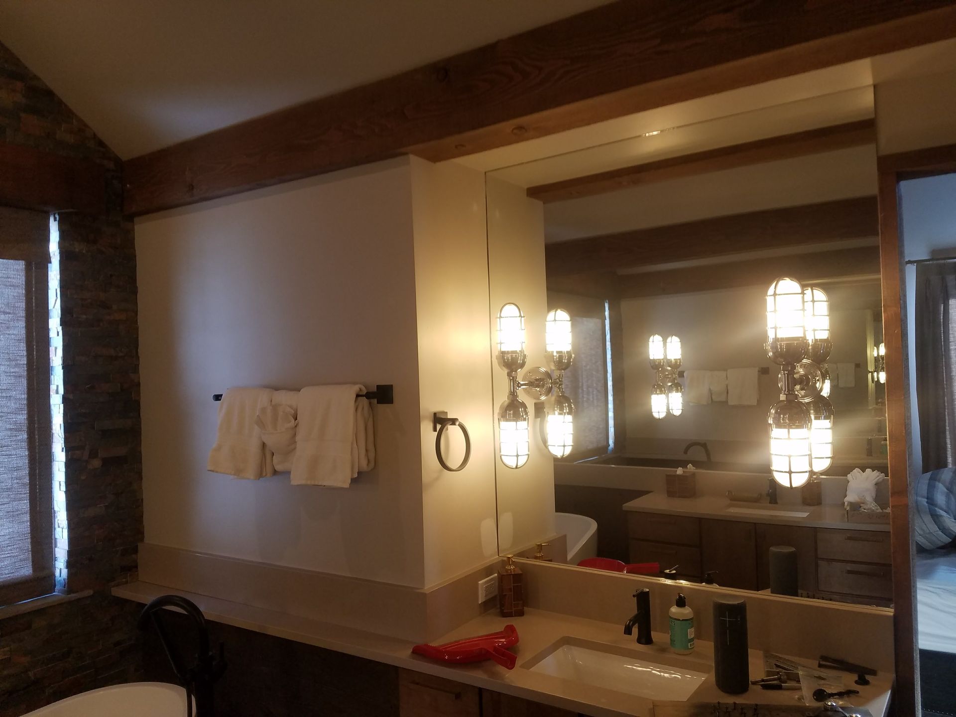 Bathroom with a large mirror reflecting the vanity, lights, and brown beams.  A towel rack is on a white wall.