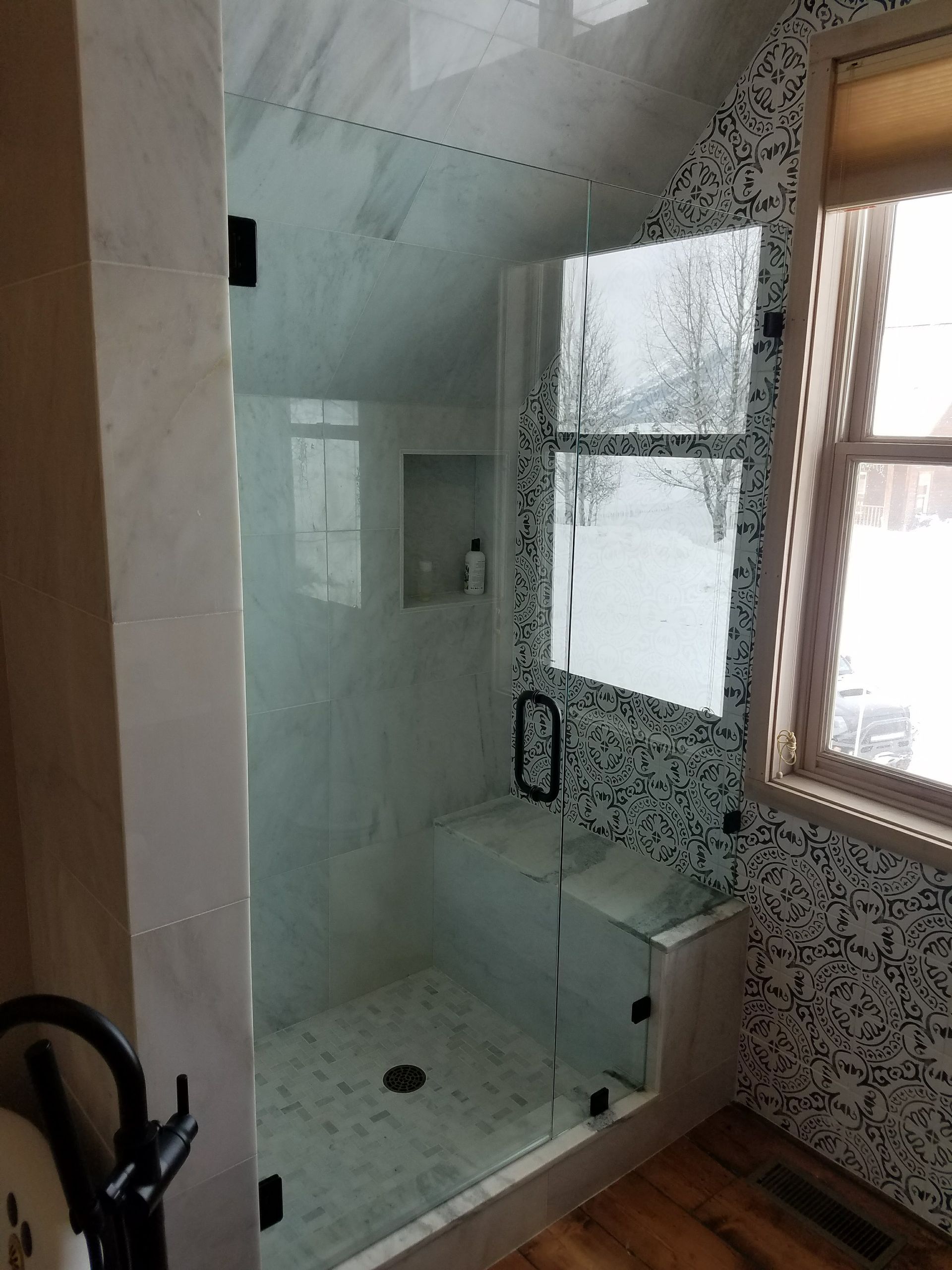 A modern shower with a glass door, marble tile, and a mosaic accent wall. There's a small bench and window to the right.