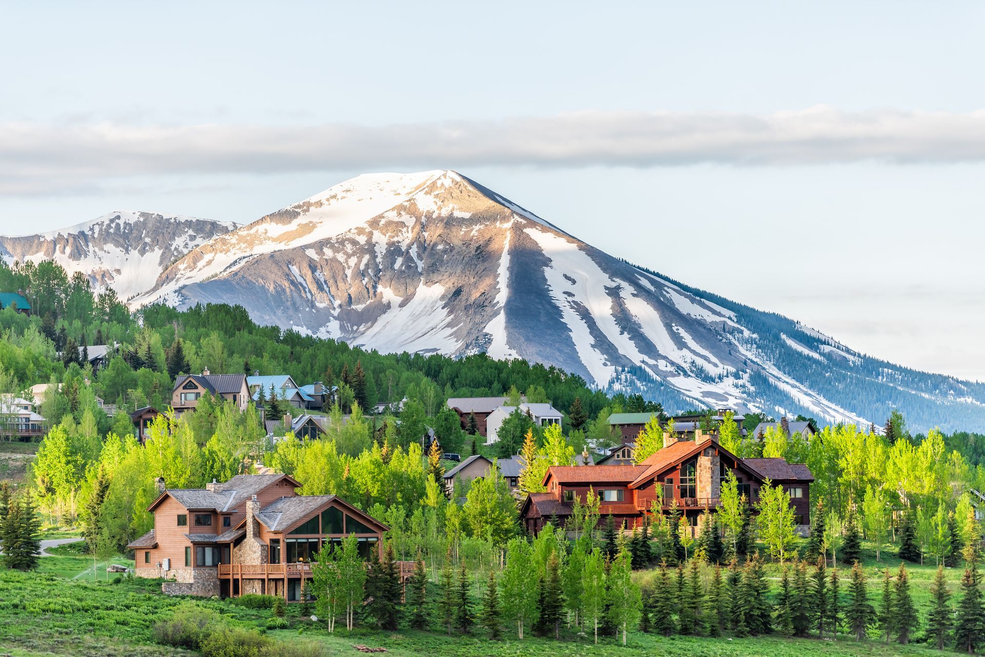 Houses nestled in trees with a snow-capped mountain backdrop. Green foliage and sunlight highlight the scene.