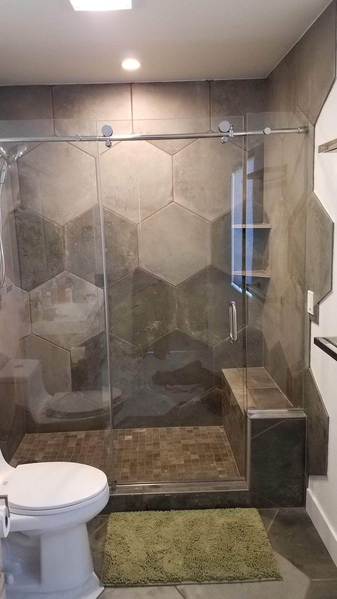 Modern bathroom with gray hexagonal tile shower and glass door; toilet and green bath mat in foreground.