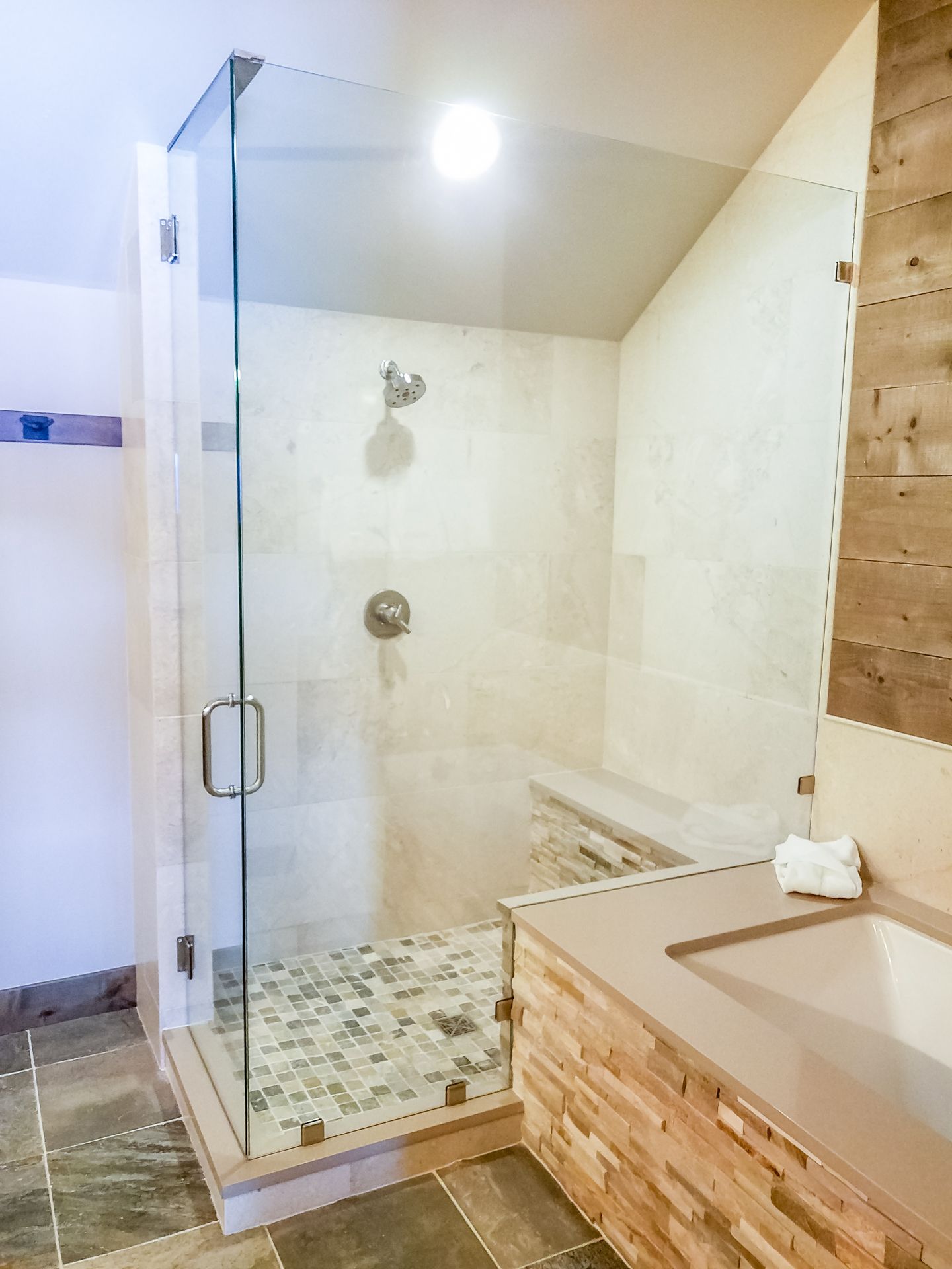 A modern bathroom with a glass-enclosed shower, stone floor, and light-colored tiled walls.