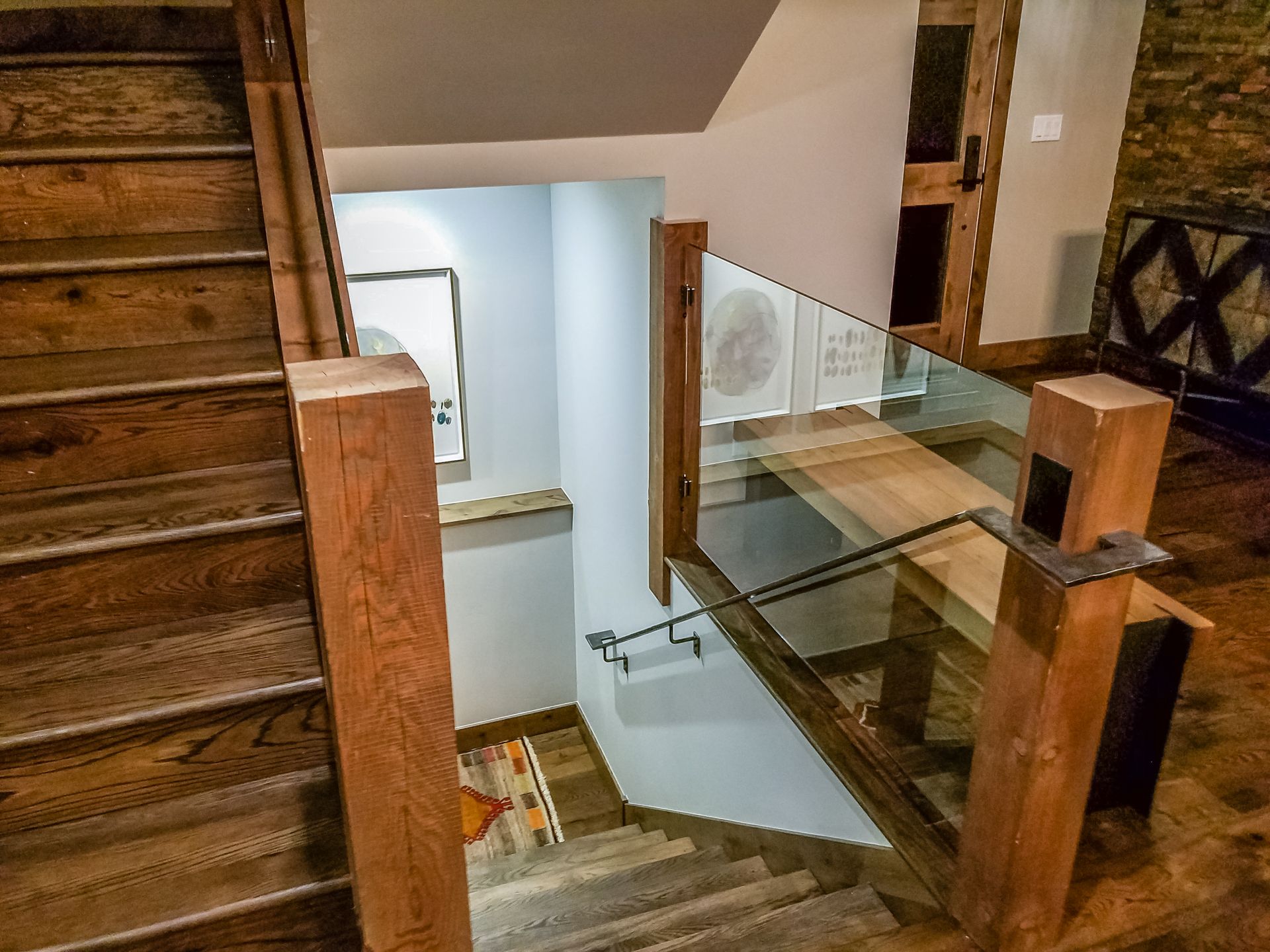 Wooden staircase descending into a lower level. Features glass railing, light gray walls, and wooden posts.