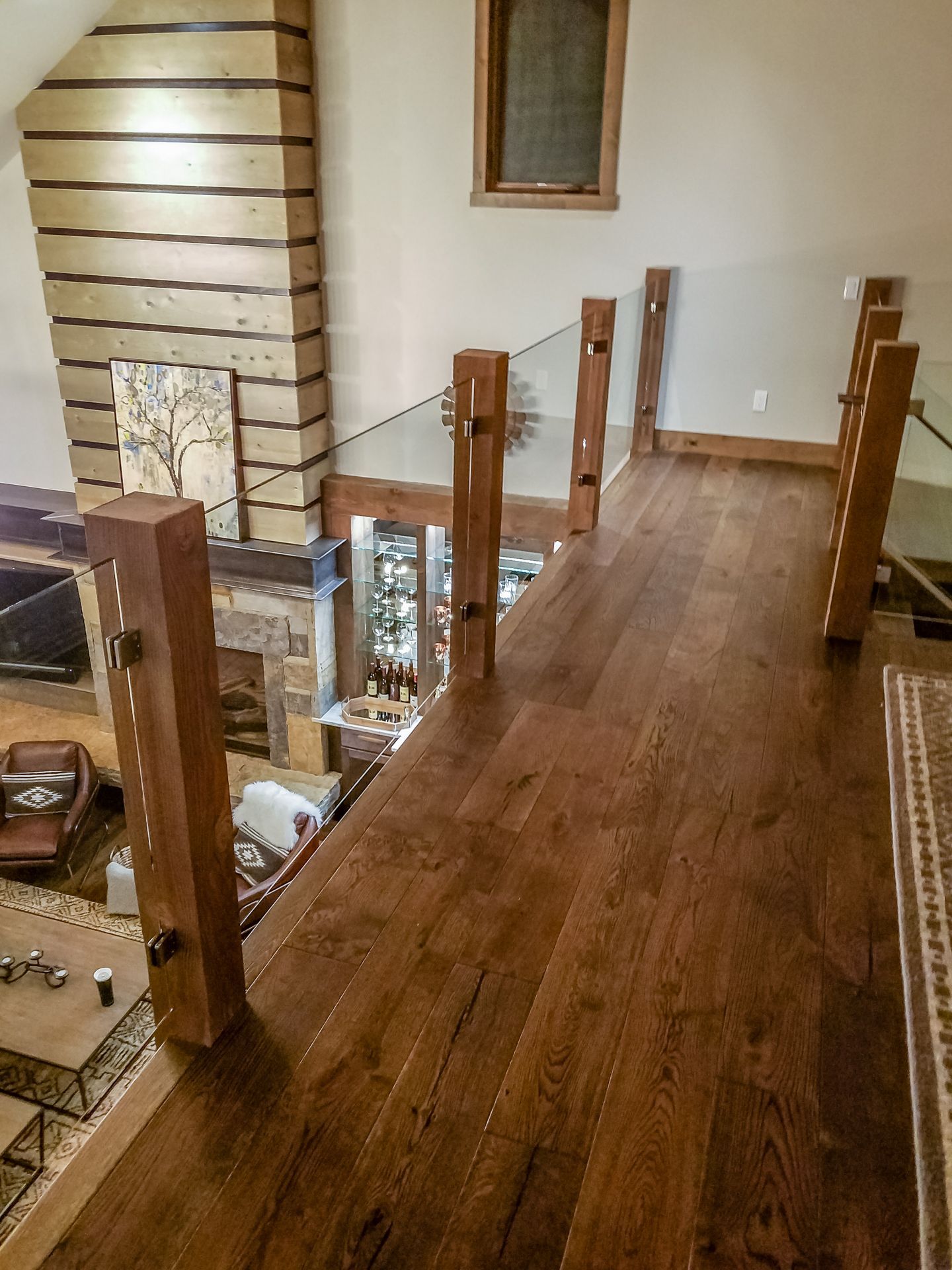 Wooden walkway with glass railing, overlooking a living room with a stone fireplace and dark furniture.