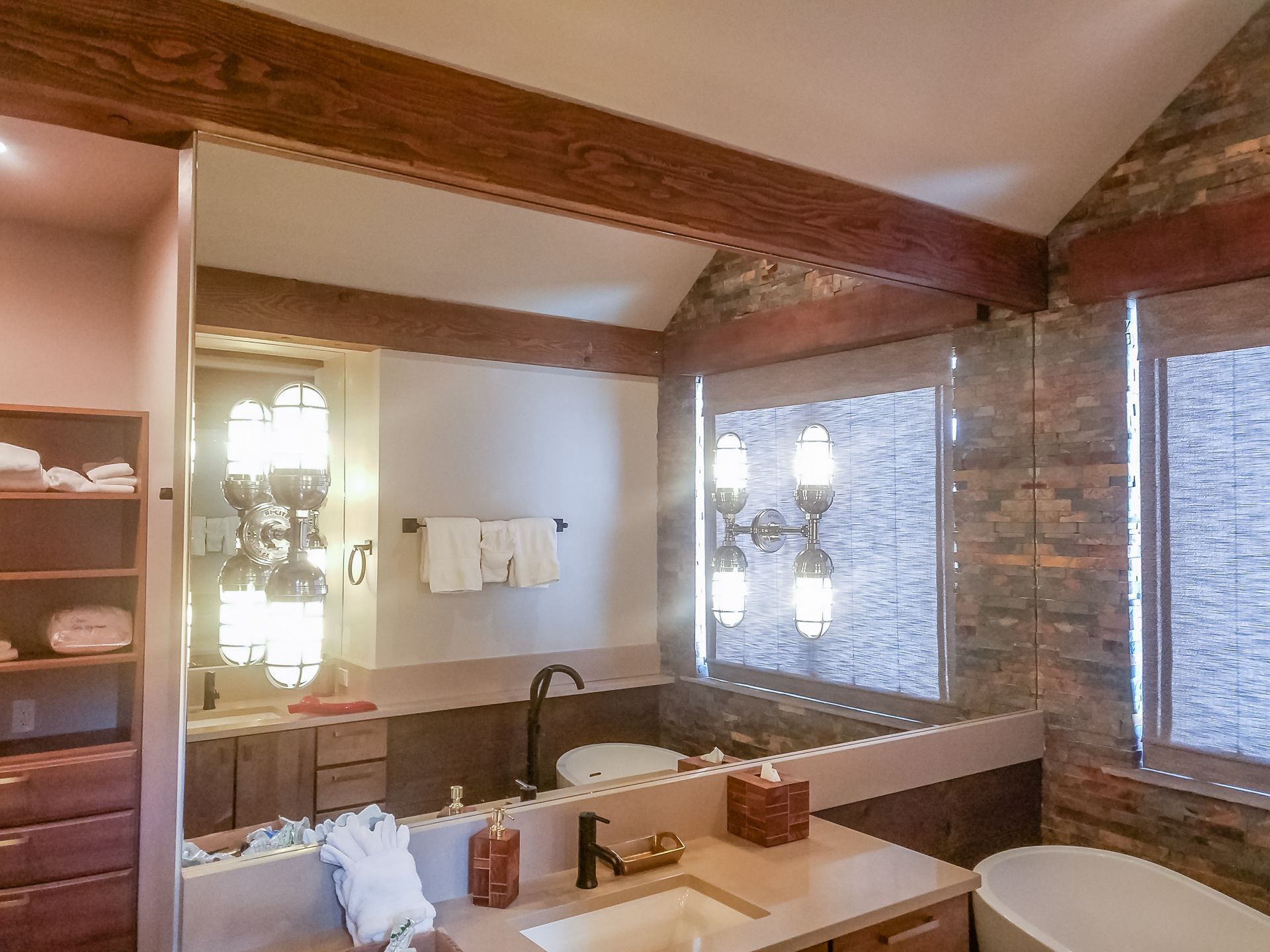 Bathroom with a large mirror reflecting a sink, lights, and a walk-in closet. Wooden beams and stone accent the walls.