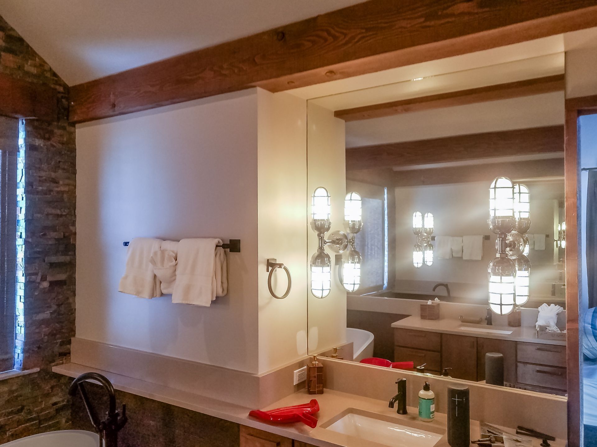 Bathroom with large mirror reflecting a vanity, towels, and stone wall. Wooden beams accent the ceiling.