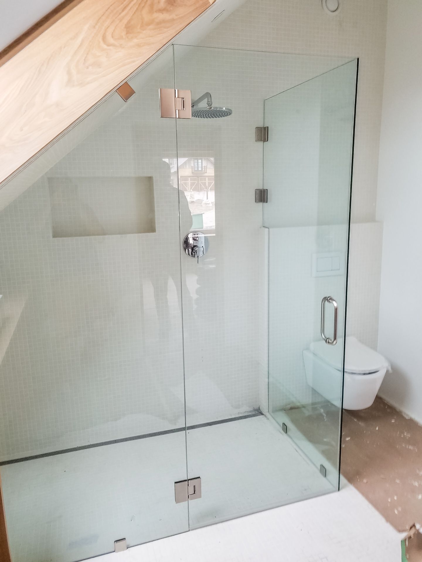 Glass shower enclosure in a bathroom, angled ceiling. Silver hardware, white tiled walls, and a toilet are visible.
