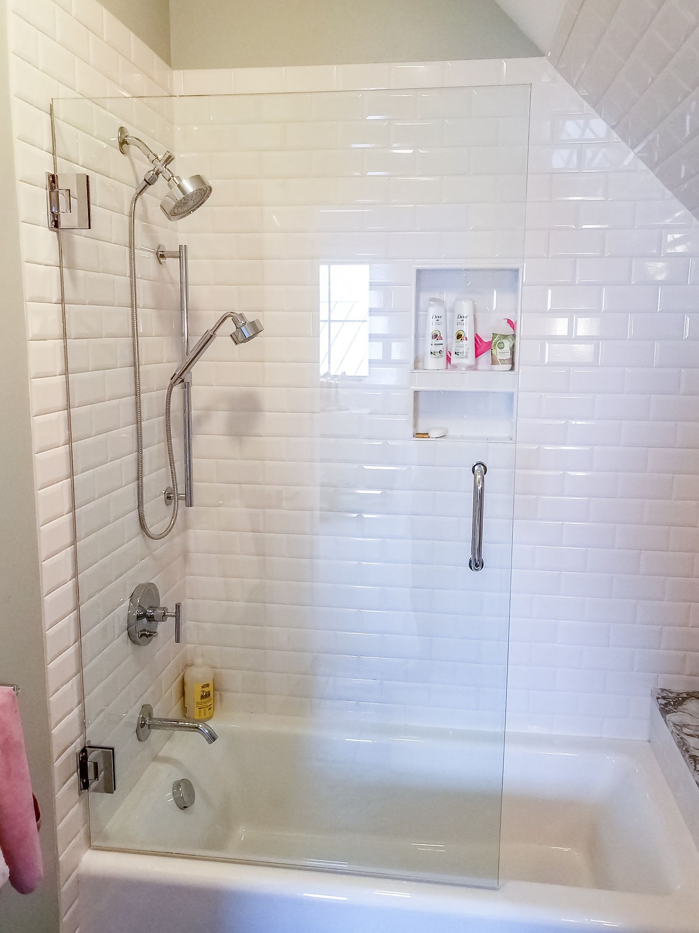 White tiled shower with glass door and built-in shelf. Includes showerhead, handheld shower, bathtub, and grab bar.