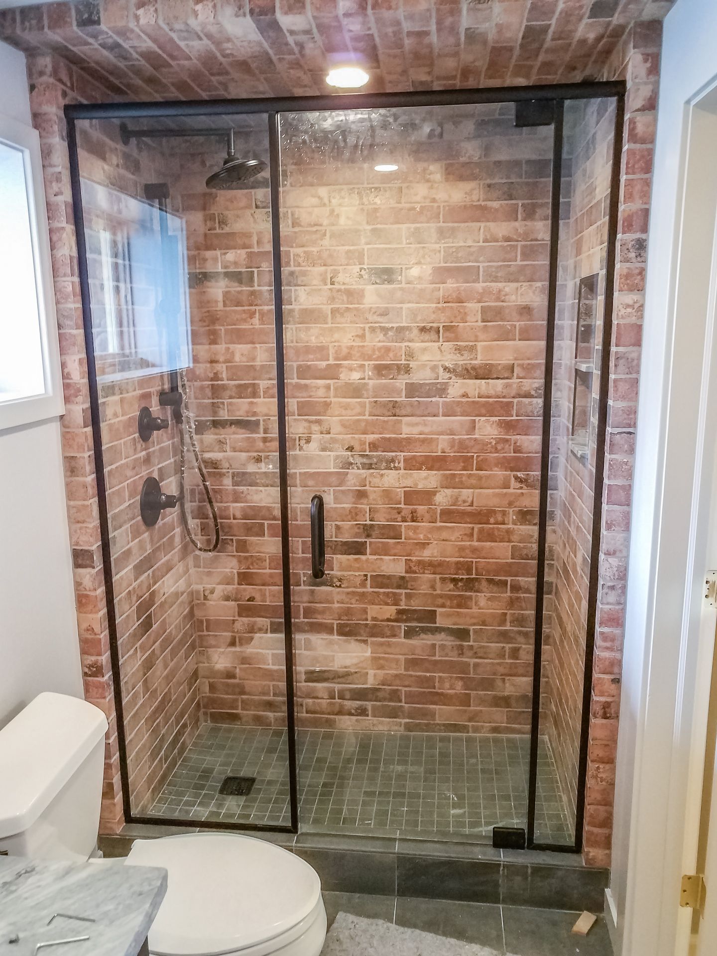 A modern shower with a brick wall interior, framed by black glass and trim. A toilet sits to the left.