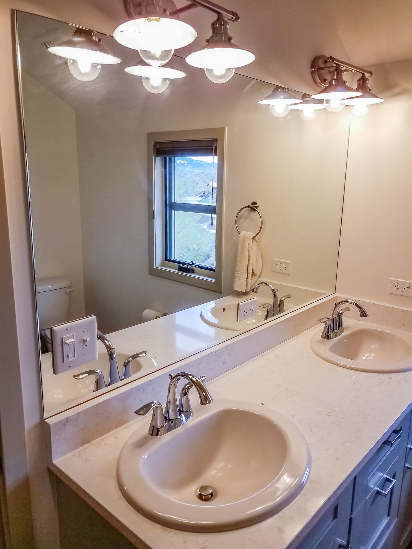 Bathroom with a large mirror above two sinks, featuring bright lighting and a small window.