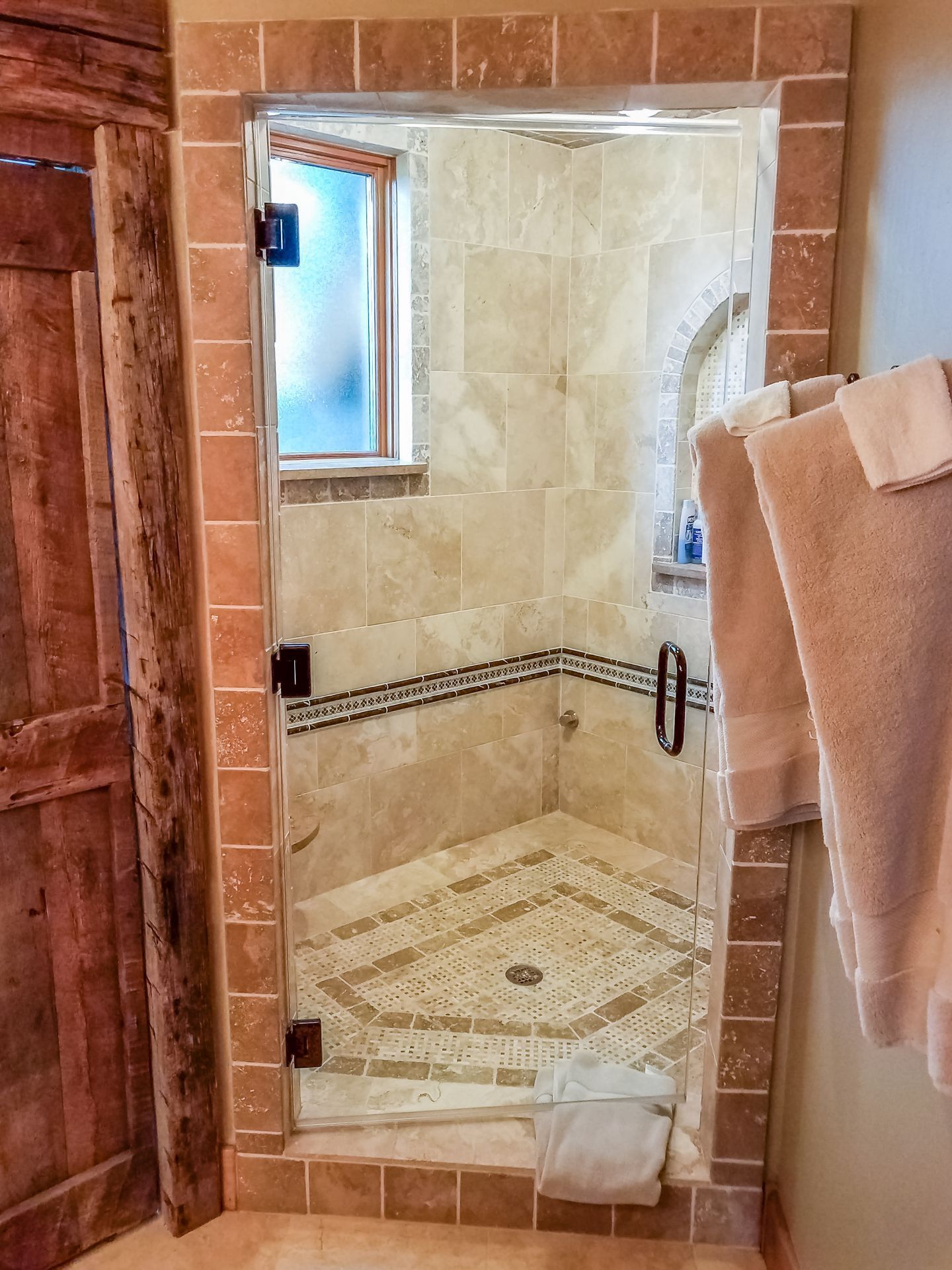 A glass shower with light-colored tile. The shower has a window and a towel rack with towels.