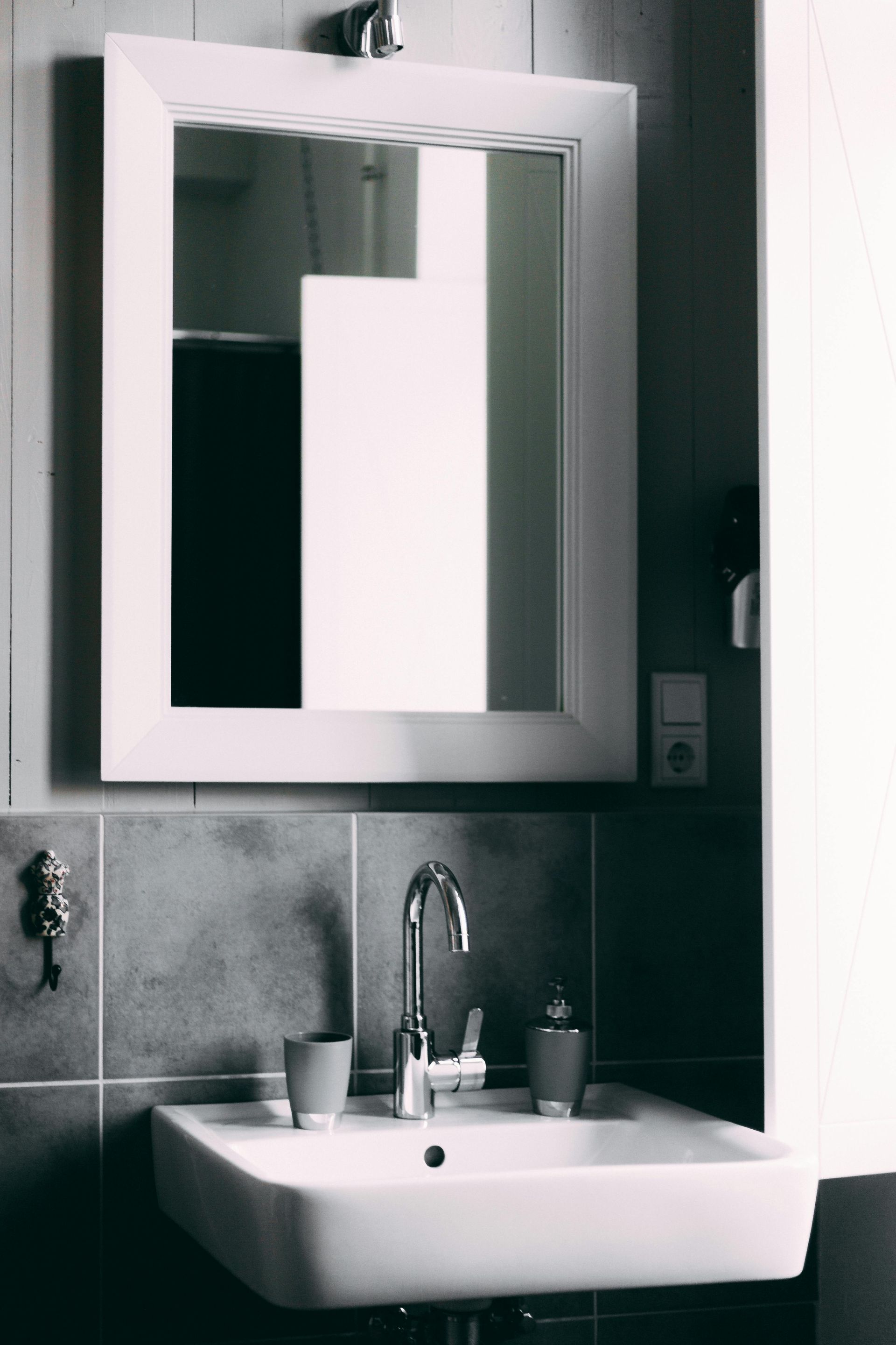 A white-framed mirror hangs above a small white sink with a chrome faucet in a tiled bathroom.