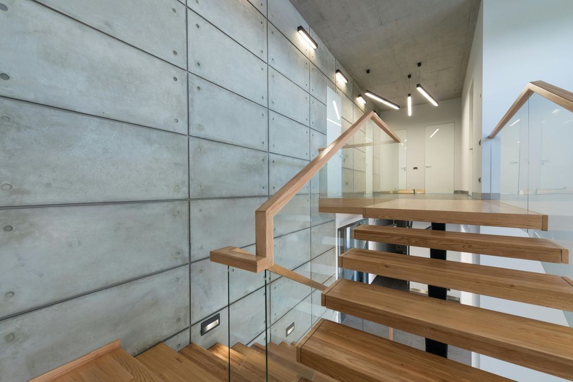 Wooden staircase with glass railing in a modern building. Concrete wall on the left; recessed lighting.